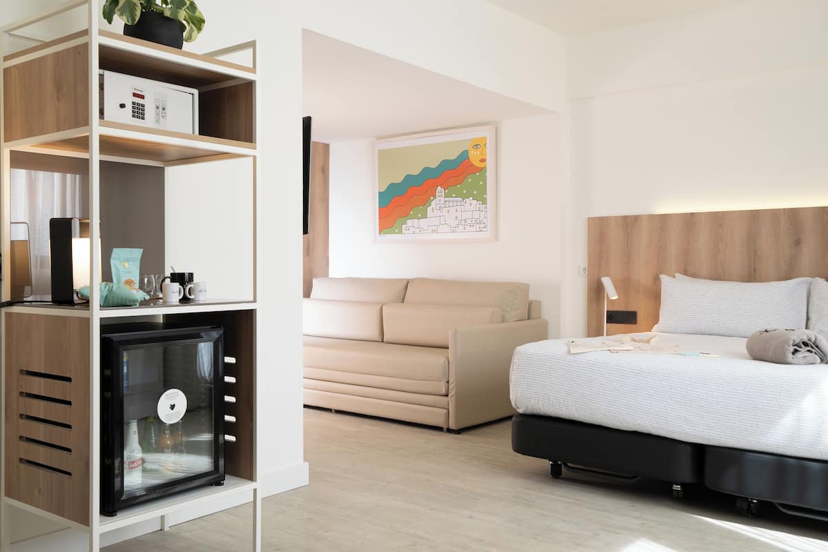 A modern hotel room with a bed, a sofa, a minibar, and a wall-mounted artwork.