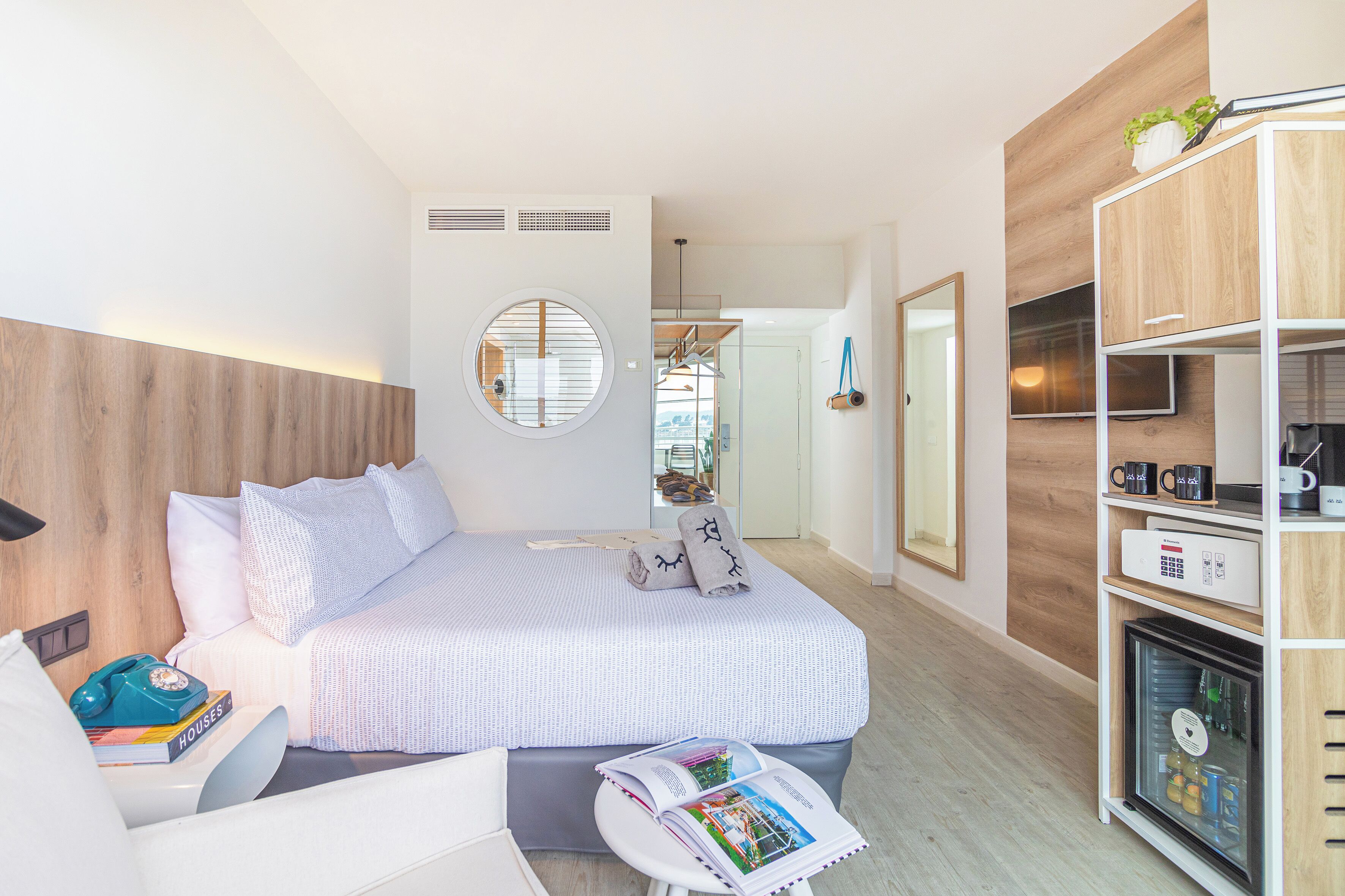 A modern hotel room with a neatly made bed, a small bedside table with a telephone and books, a magazine, and a minibar.
