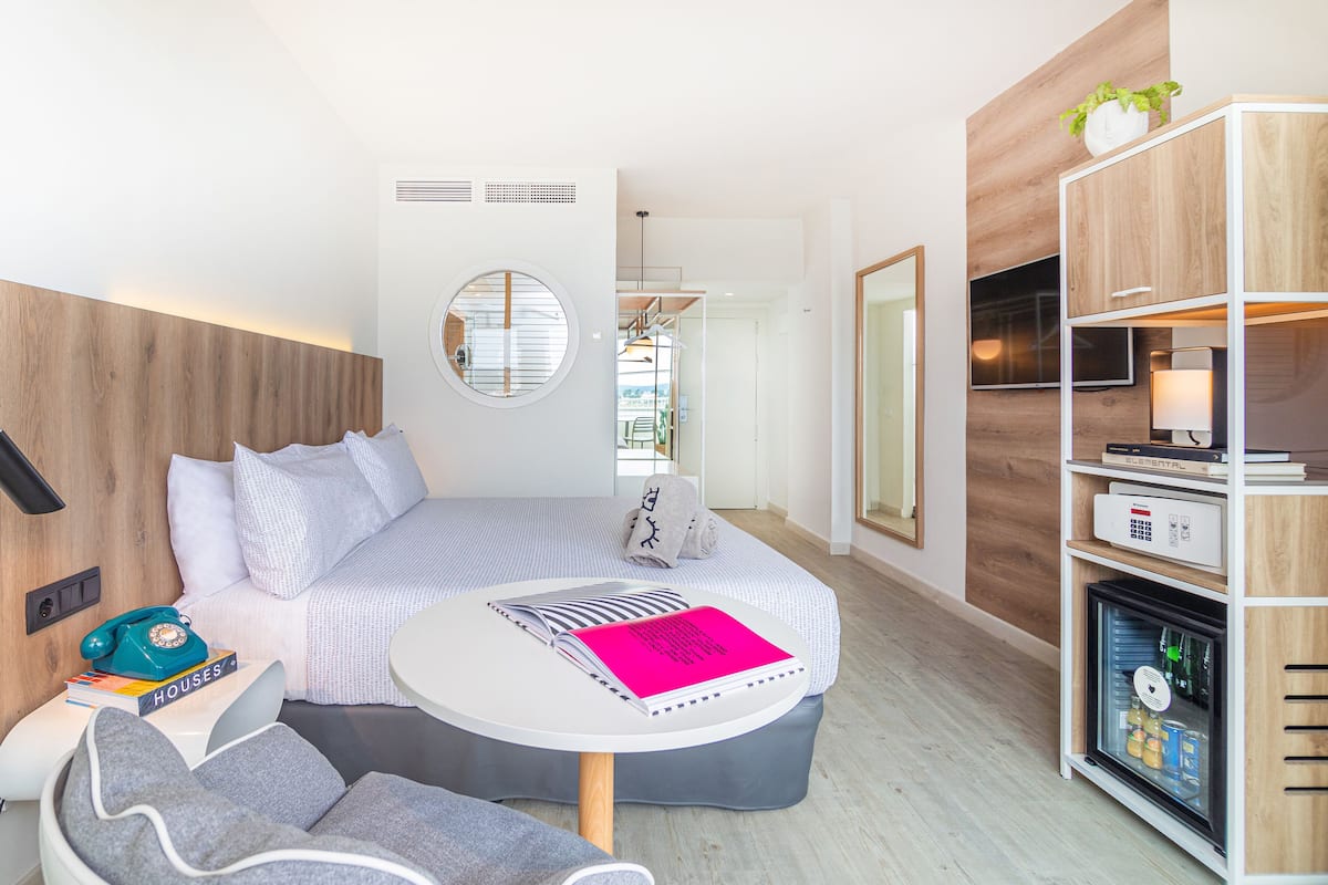A compact hotel room with a bed, bedside table, and a small seating area. There is a minibar and a wall-mounted TV.