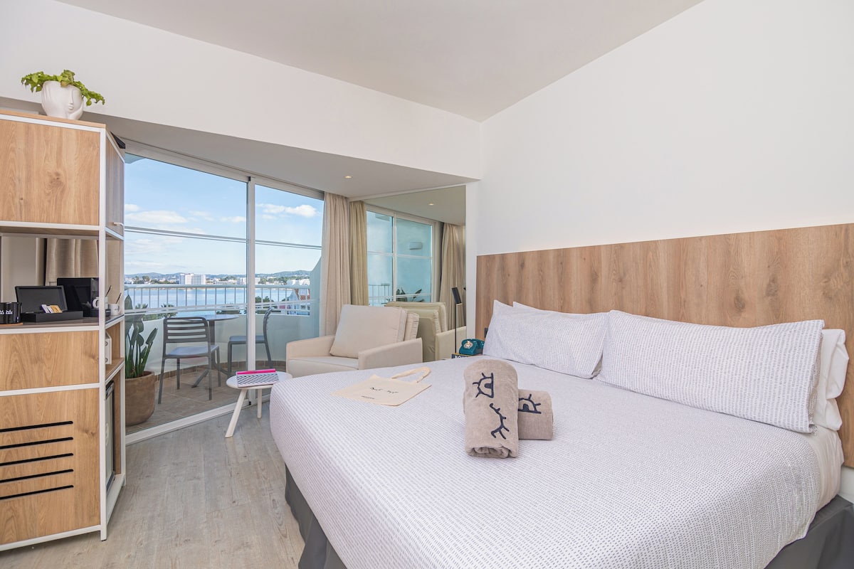 A modern hotel room with a large bed, a desk, a chair, and a balcony with a view.
