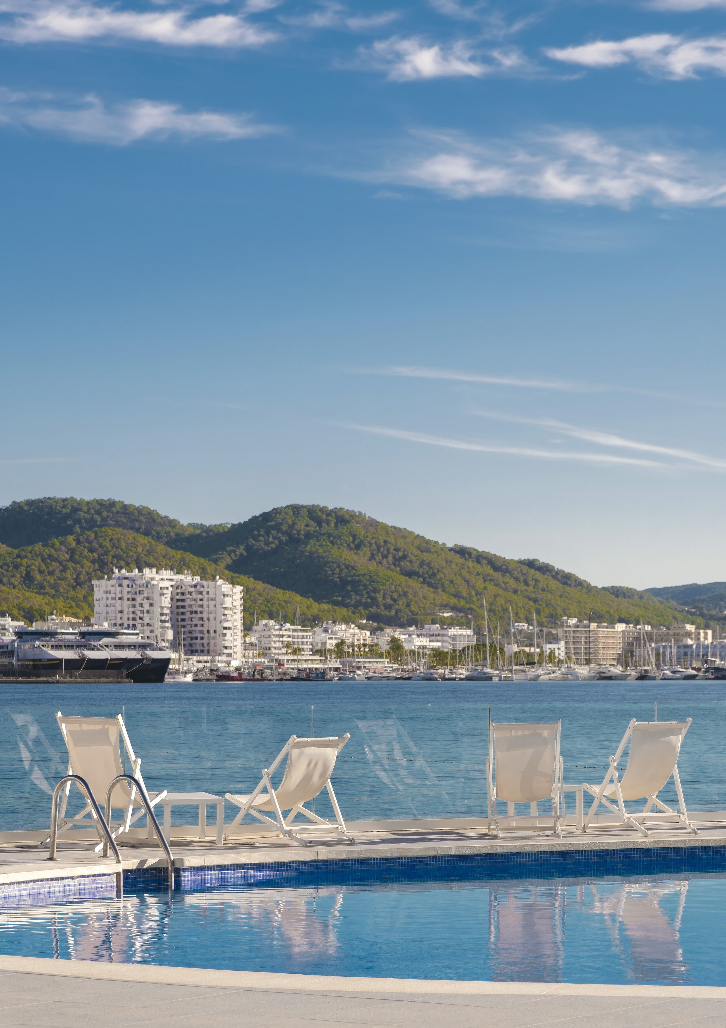 Vista de la piscina con sillones de descanso, cielo azul despejado y un puerto deportivo con barcos y edificios al fondo.