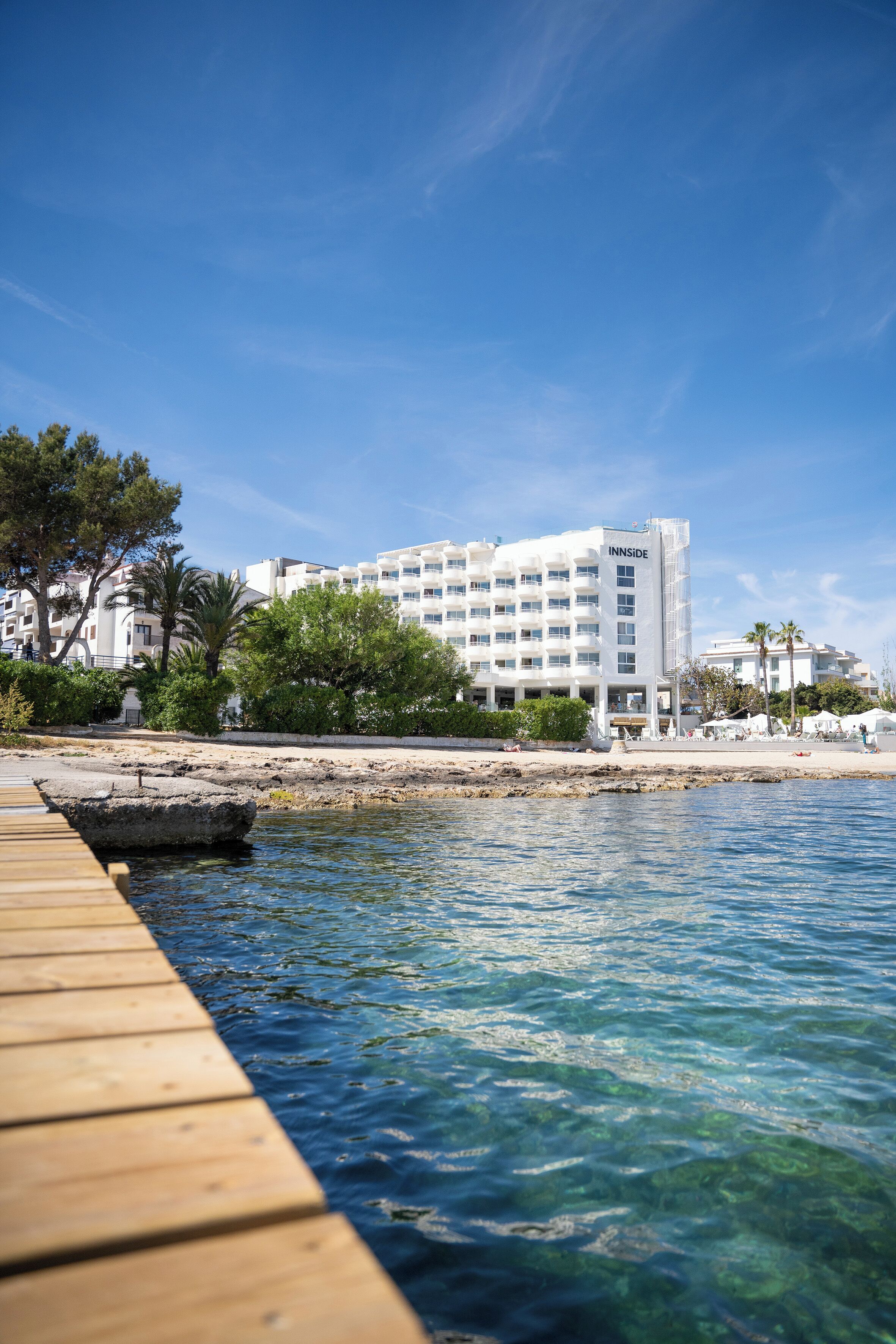 Un hotel frente al agua con un muelle de madera, agua azul cristalina y un edificio blanco con balcones.