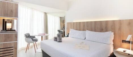 A modern hotel room with a large bed, a desk, a chair, and a wardrobe.