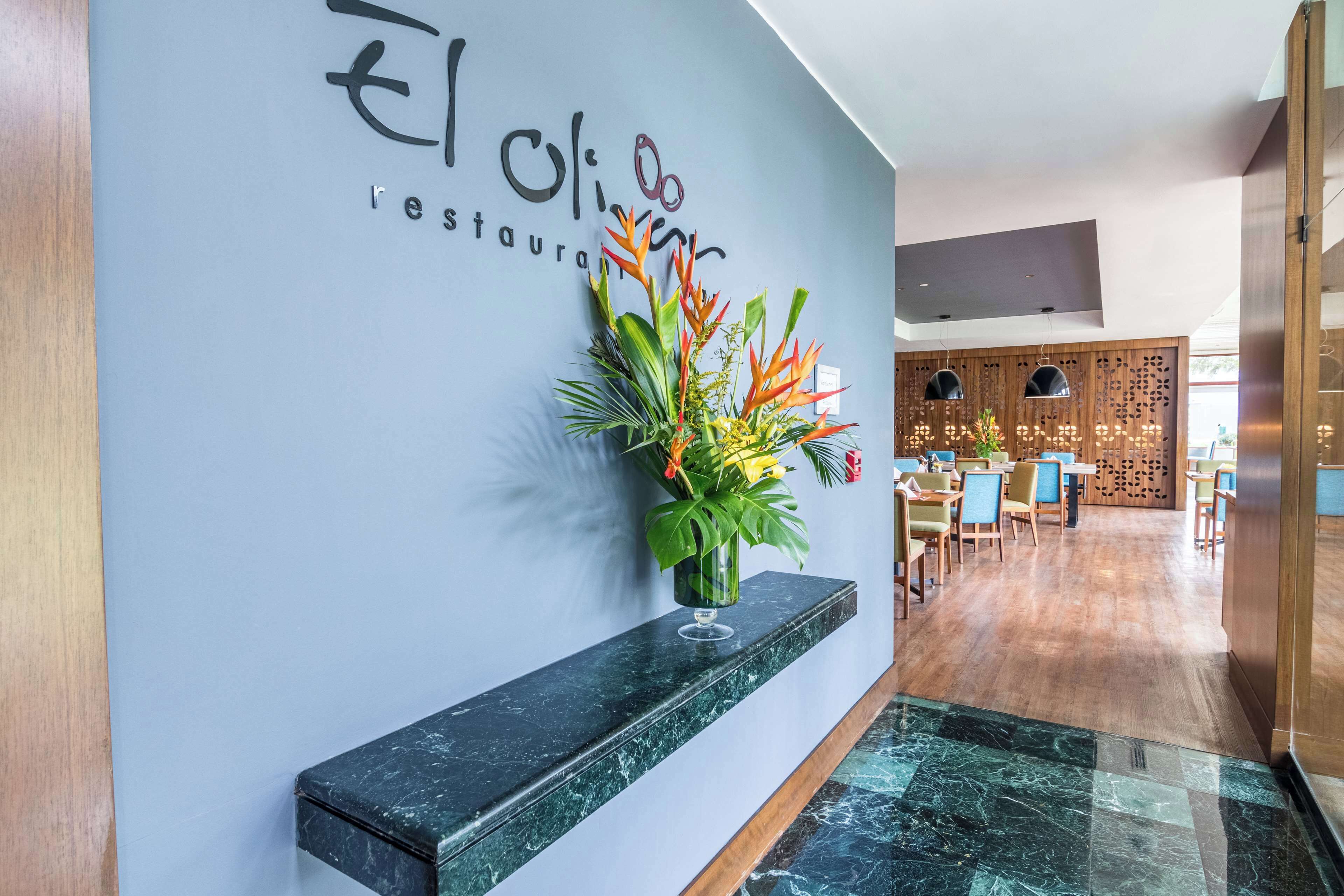 Interior moderno de restaurante con una recepción de mármol, una planta montada en la pared y una zona de estar con mesas y sillas de madera.