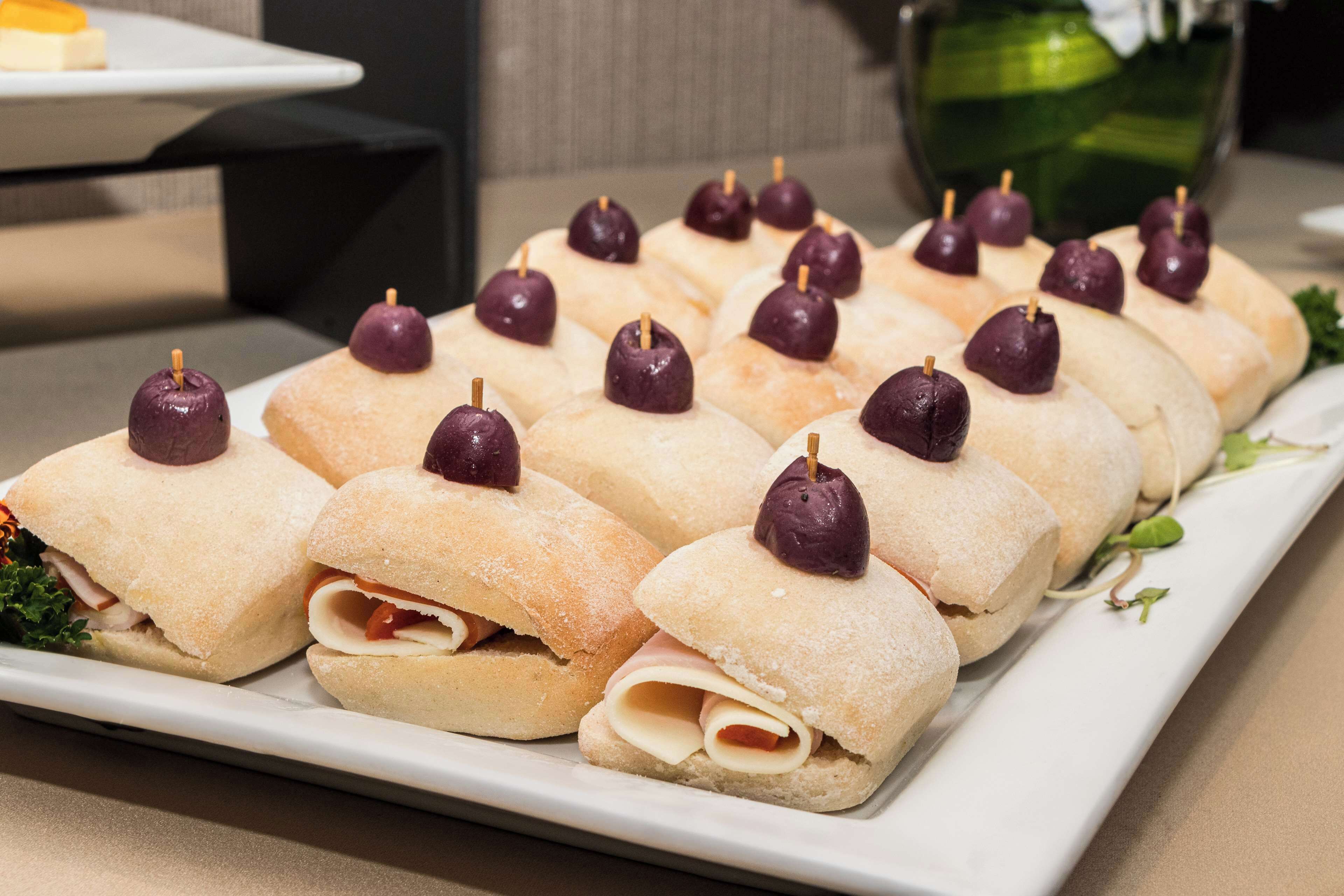 A white rectangular plate with several sandwiches, each topped with a purple cherry and a toothpick.