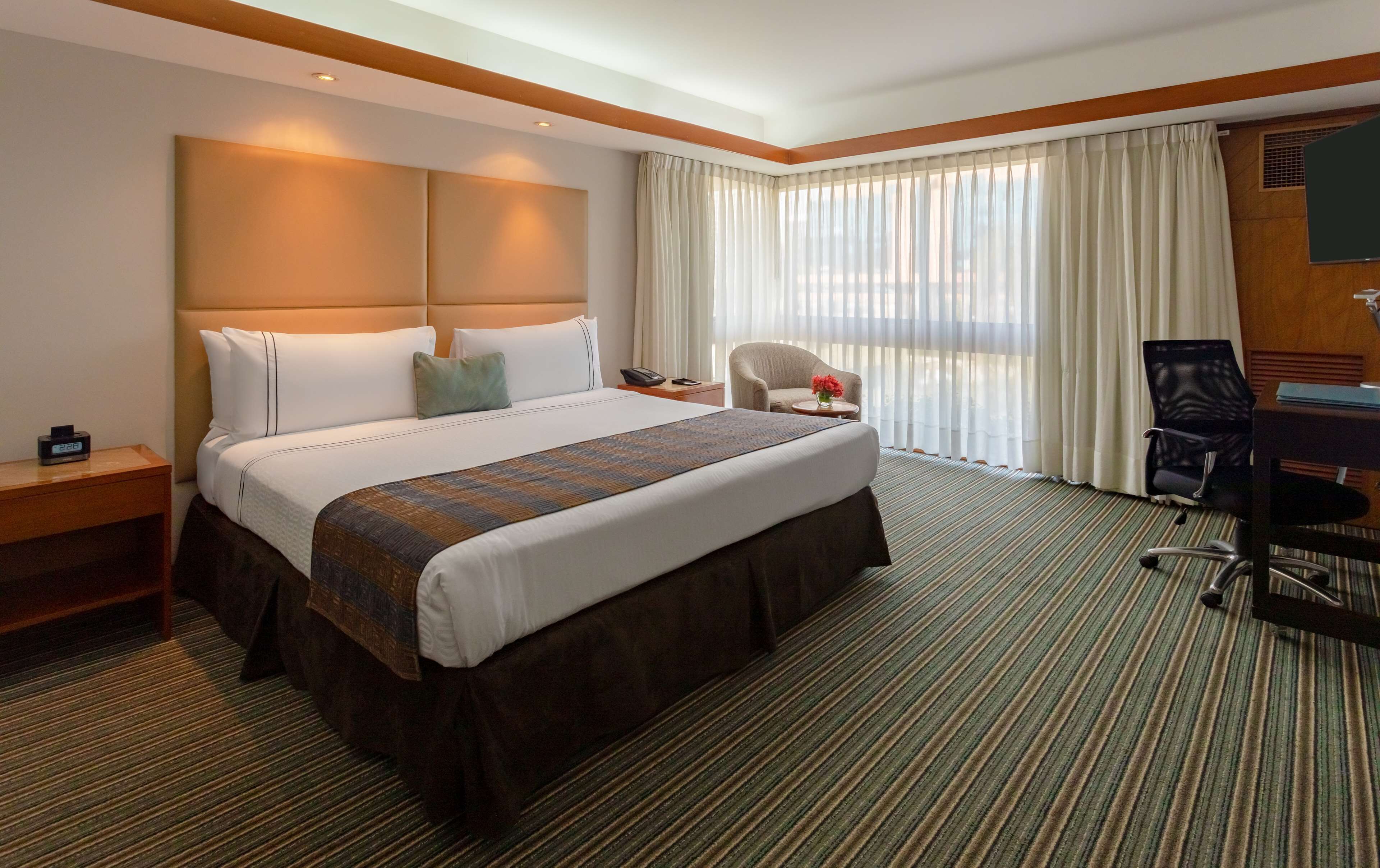 Habitación de hotel con una cama grande, un escritorio, una silla y vista a la ciudad por la ventana.