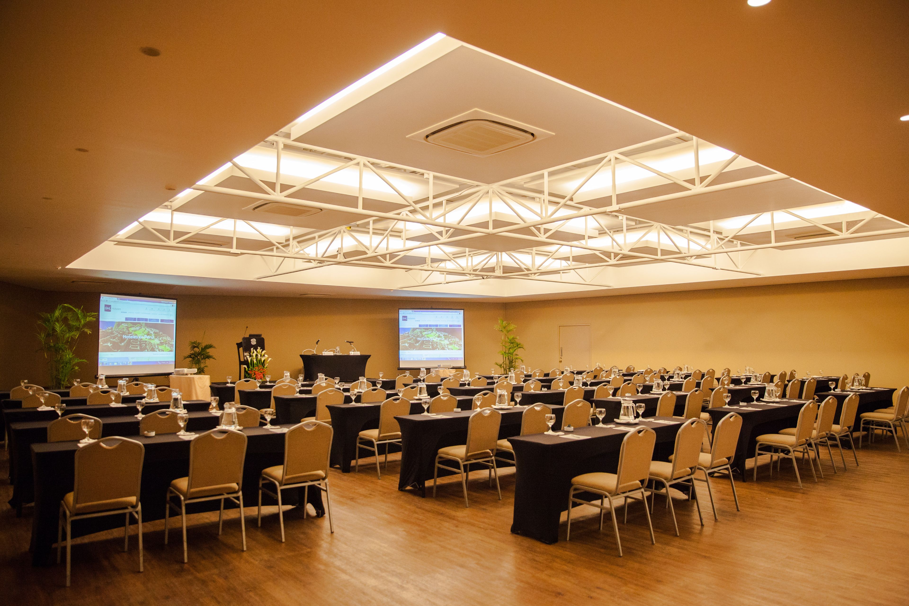 Un salón de conferencias con varias mesas y sillas, equipado con proyectores y un atril.