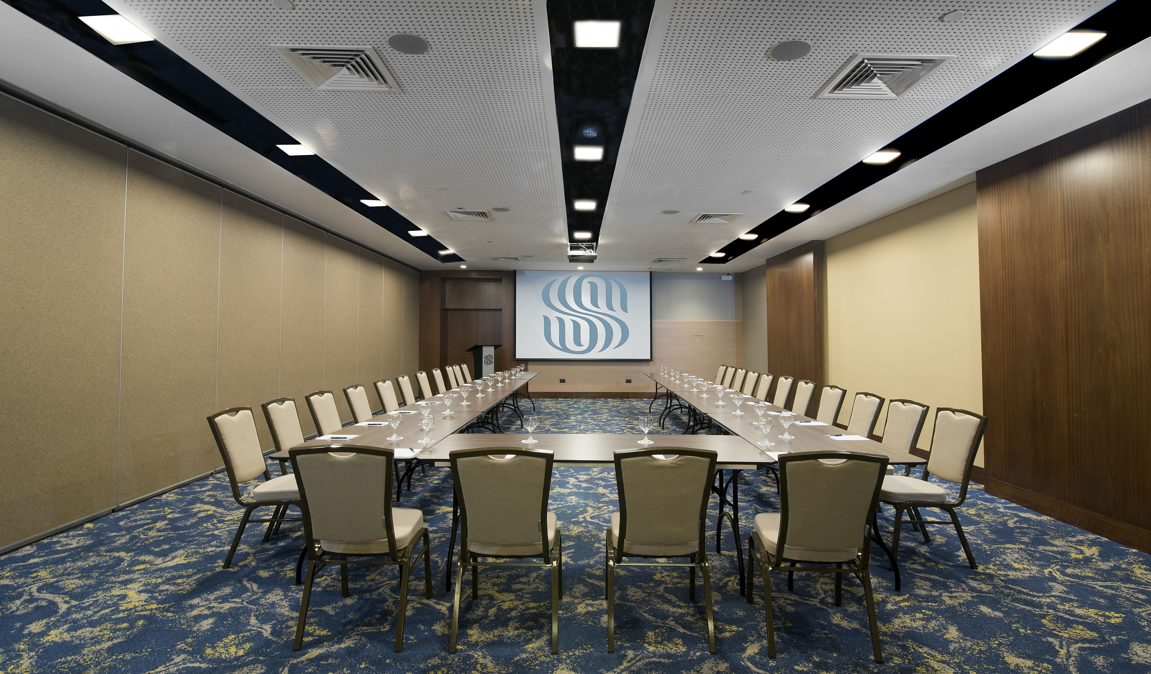 A conference room with a long table, chairs, a podium, and a screen with a logo.