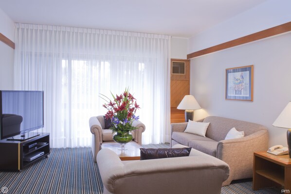 A hotel room with a flat-screen TV, two sofas, a coffee table with a vase of flowers, a lamp, and a painting on the wall.