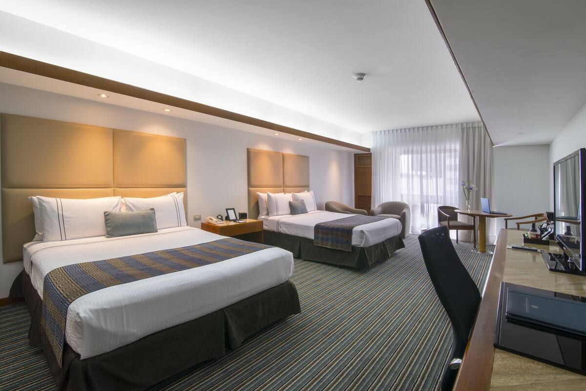 Quarto de hotel com duas camas, uma escrivaninha, uma cadeira e uma televisão.