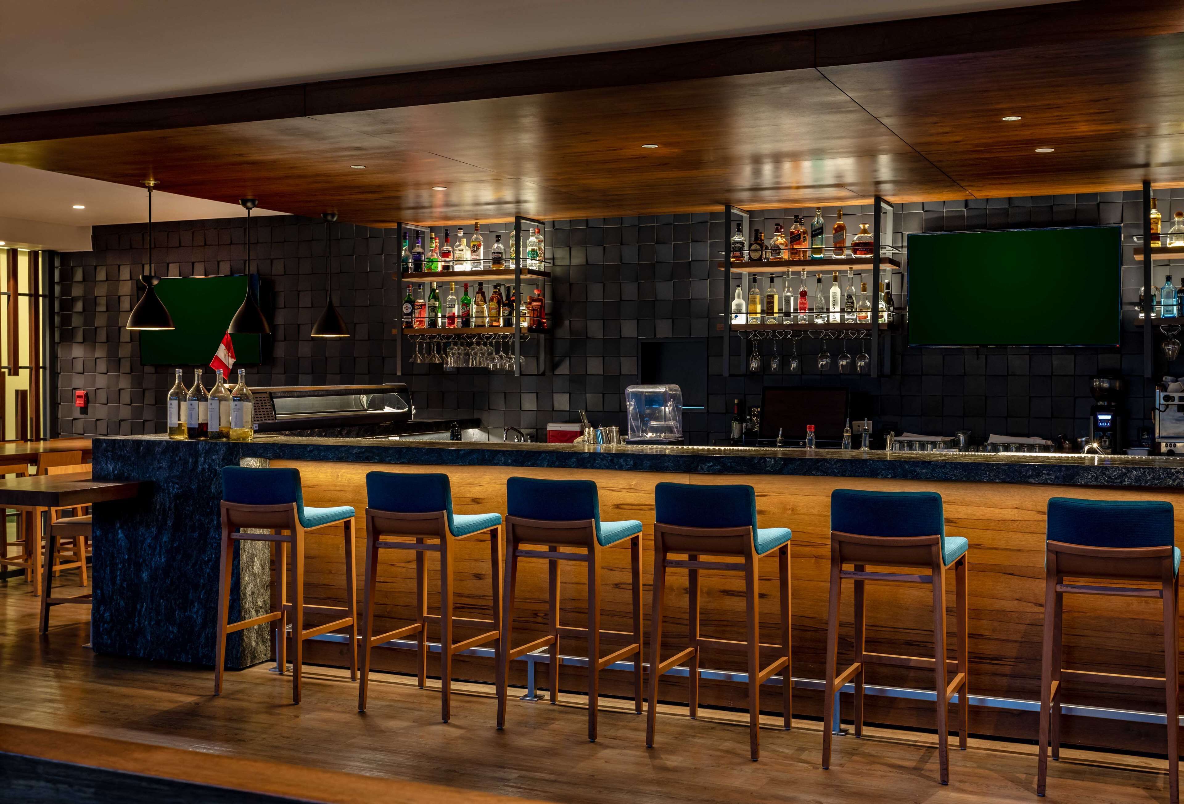 Een bar met een donkere, getextureerde wand, een houten toog en meerdere flessenkasten. Er staan barstoelen bij en een televisie hangt aan de wand.