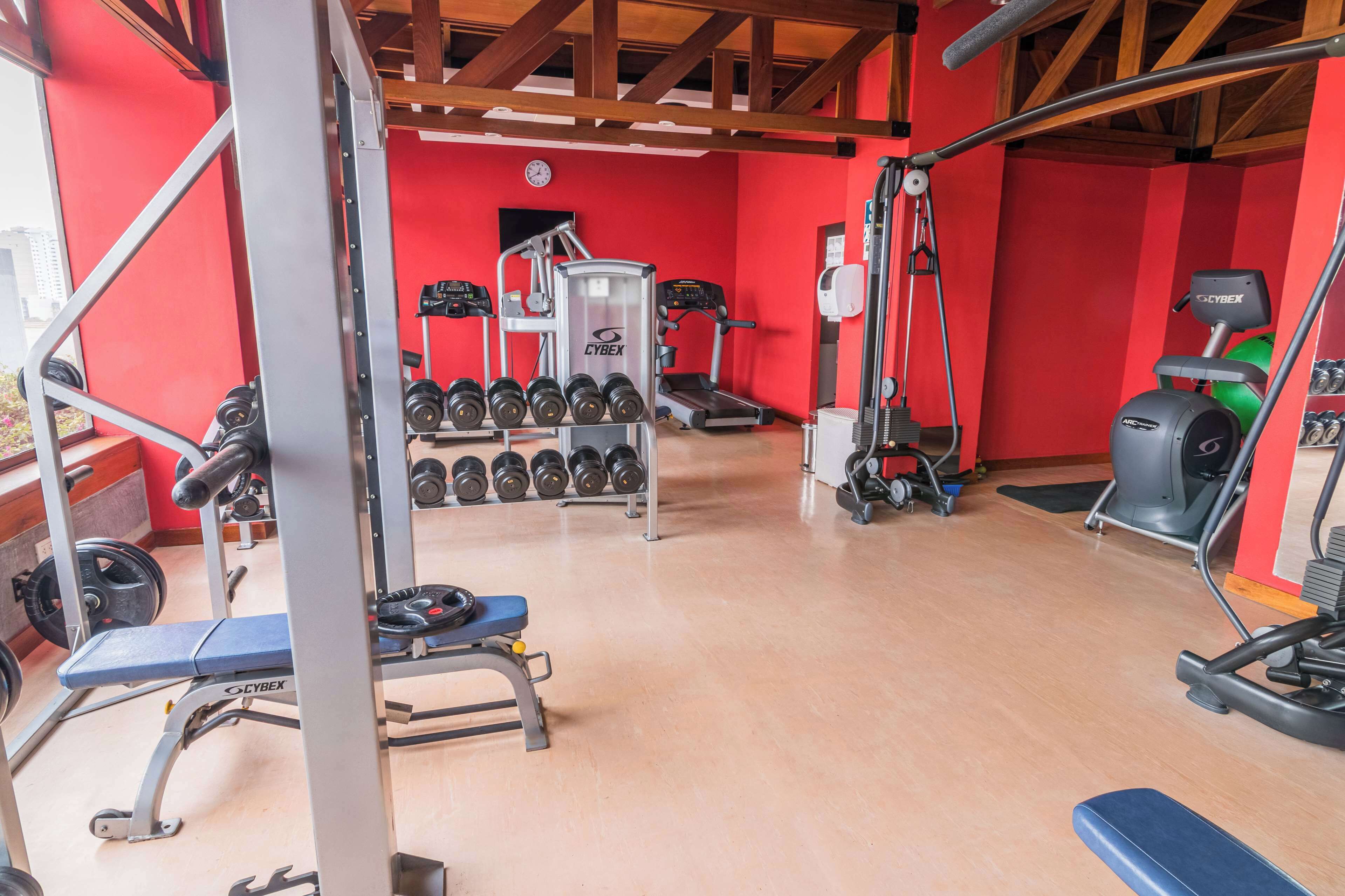 Ein Fitnessstudio mit roten Wänden, verschiedenen Trainingsgeräten und einer Holzzieldecke.