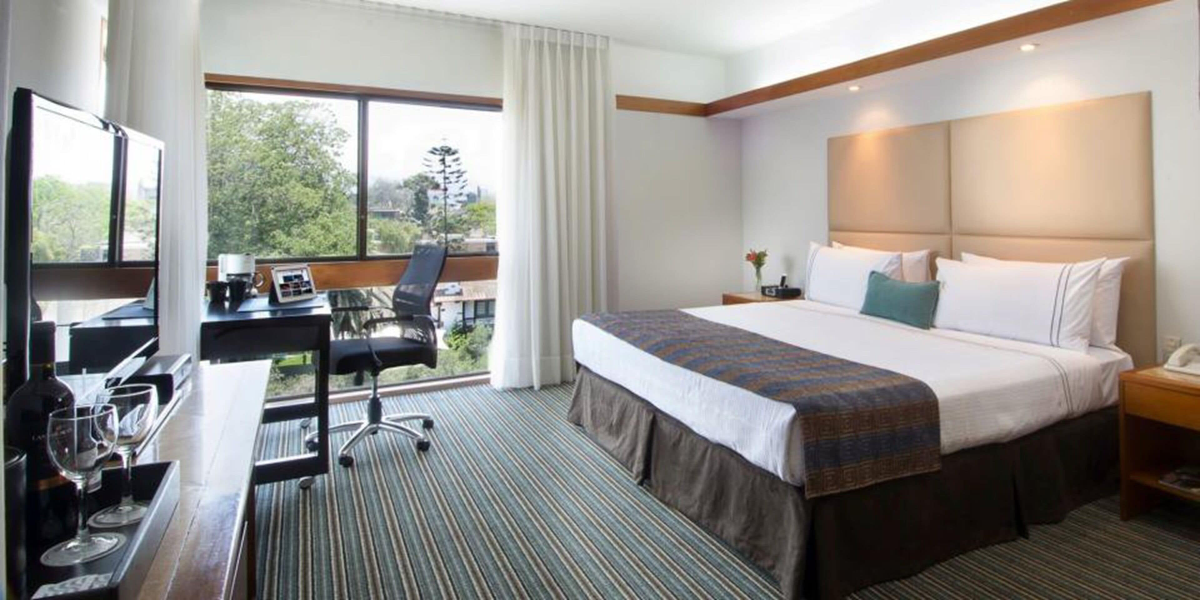 Une chambre d’hôtel avec un grand lit, un bureau avec une chaise, une fenêtre avec des rideaux et une vue sur les arbres et les bâtiments environnants.