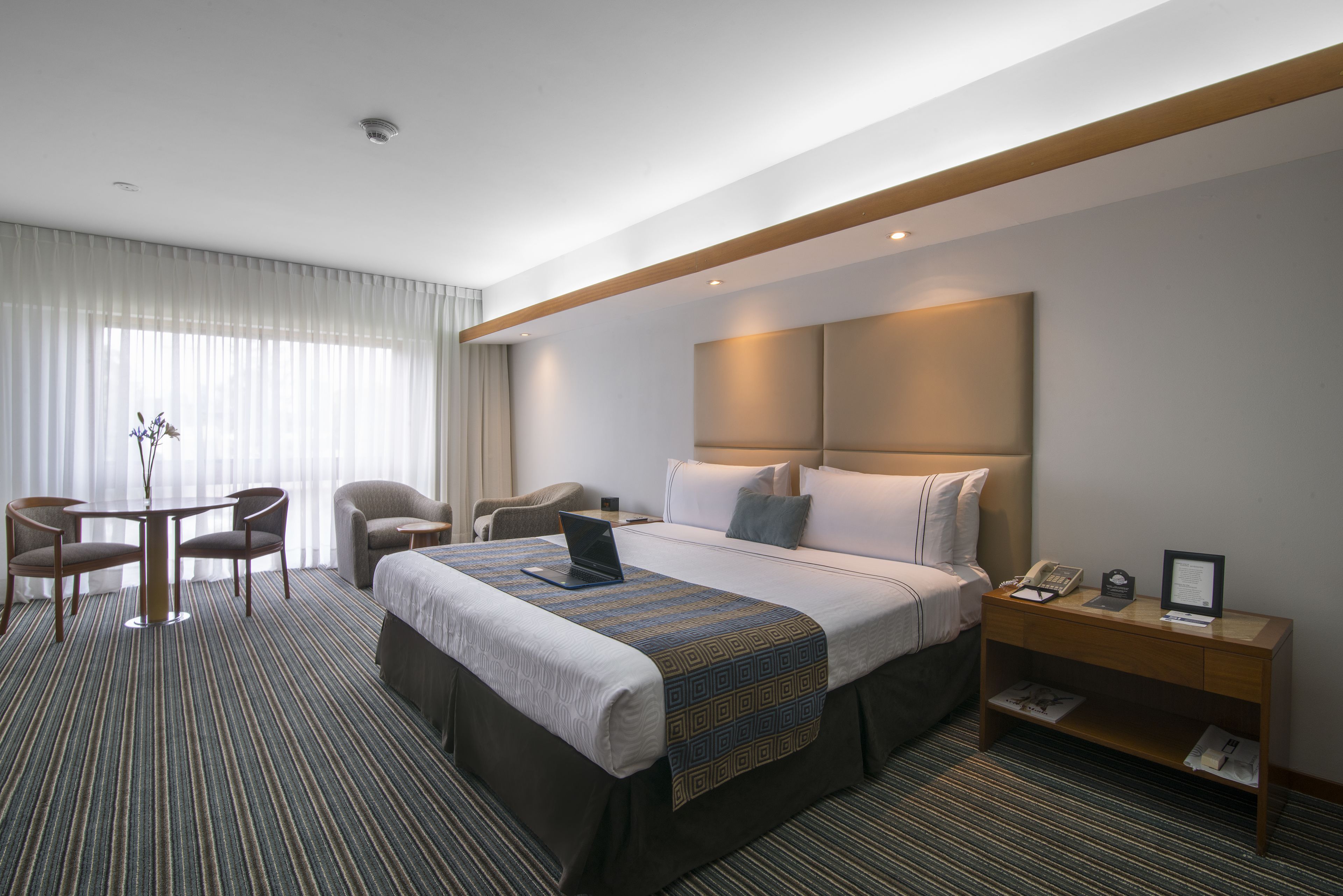 Habitación de hotel con una cama grande, un escritorio, una silla, una computadora portátil y una mesita de noche con un teléfono y un marco de fotos.