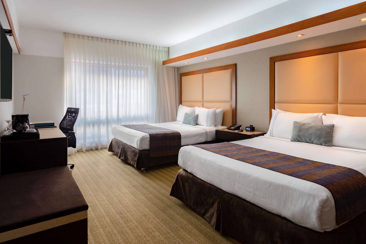 Quarto de hotel com duas camas, uma escrivaninha, uma cadeira e uma janela grande com cortinas.