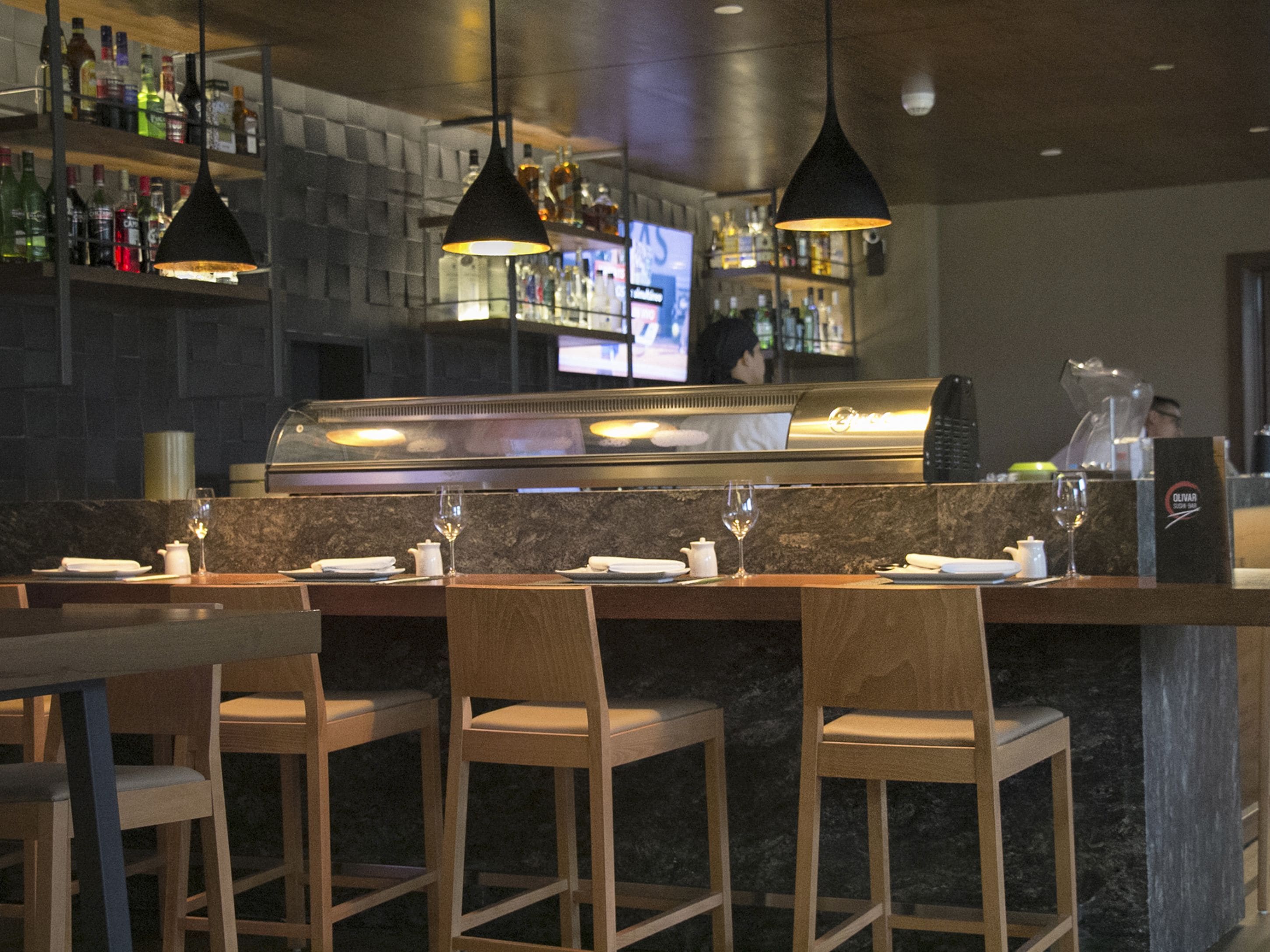 Uma área de bar com cadeiras de madeira, um balcão com vitrine de vidro e luminárias pendentes.