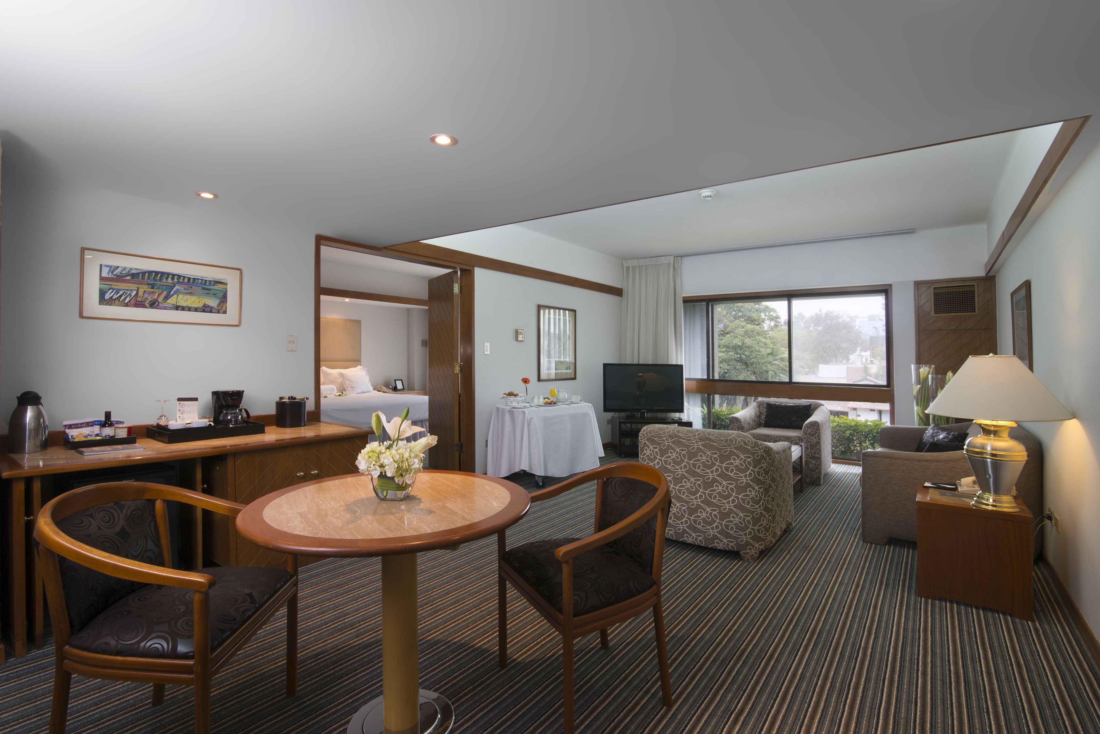 A hotel room with a dining area, a sofa, a TV, and a window with a view of trees.