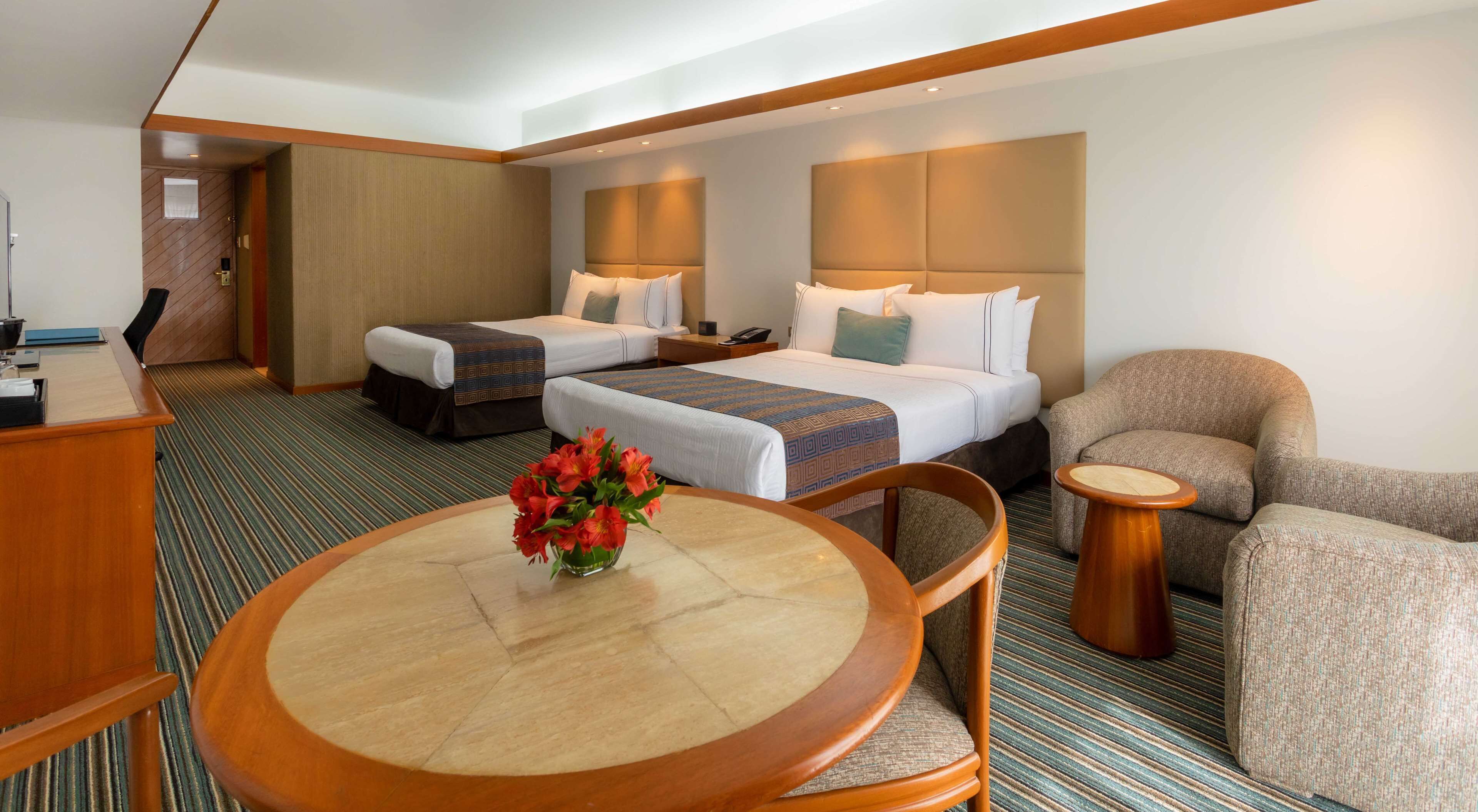 Habitación de hotel con dos camas, un escritorio, una silla, una mesa redonda y un jarrón con flores.