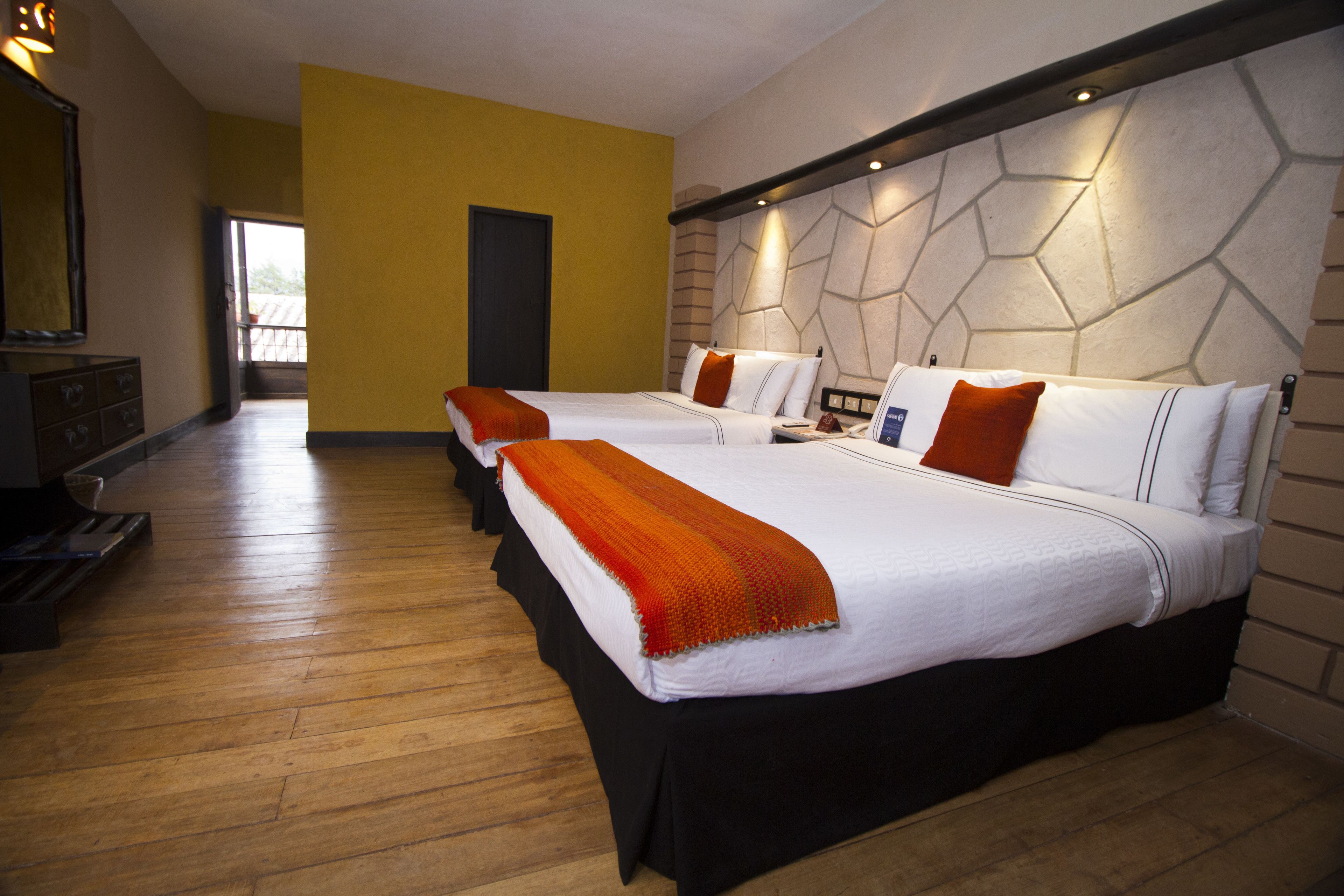 A hotel room with three beds, wooden flooring, a modern wall design, and a large mirror.