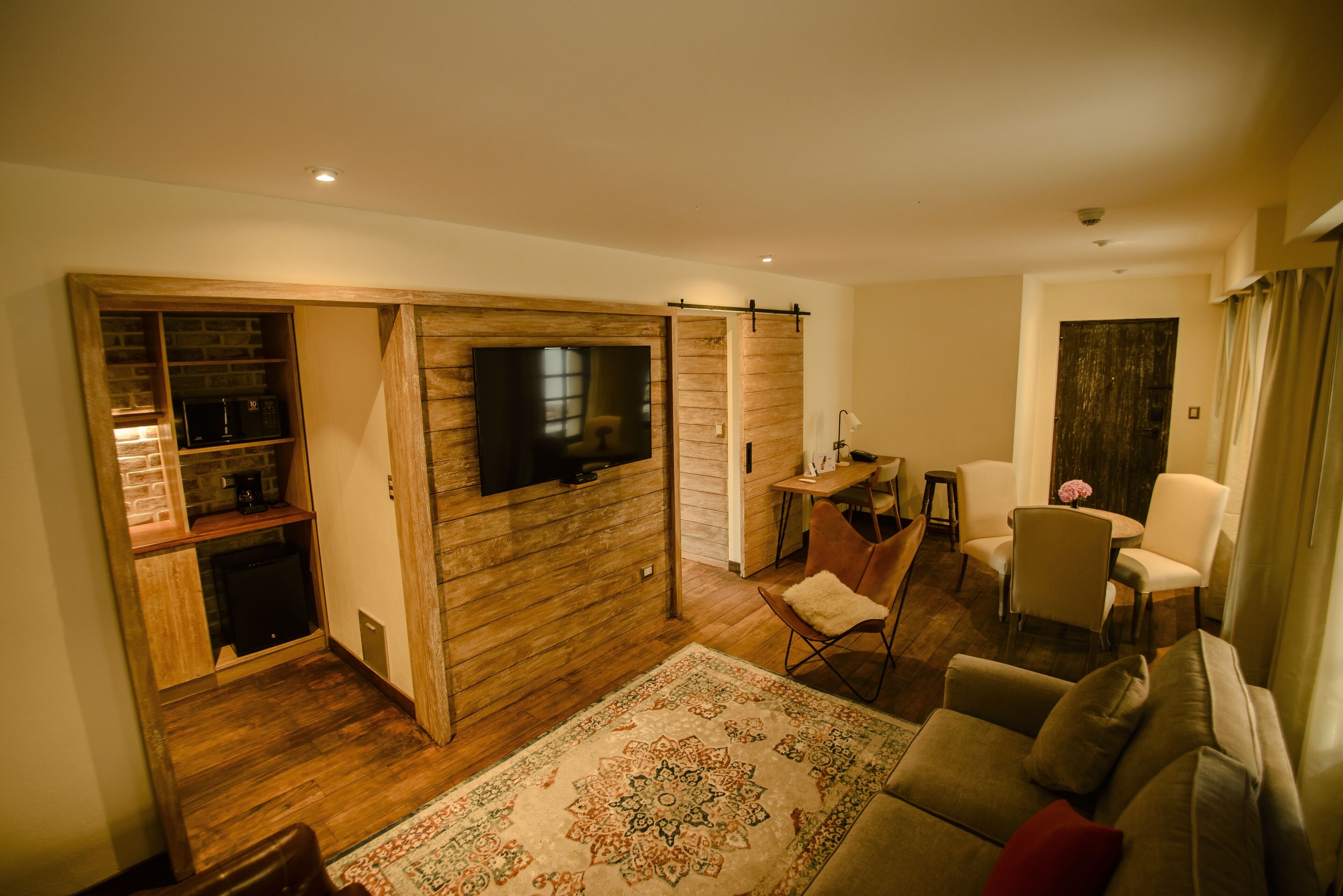 A living room with a wooden floor, a large TV mounted on a wall, a sofa, a coffee table, and a small dining area.