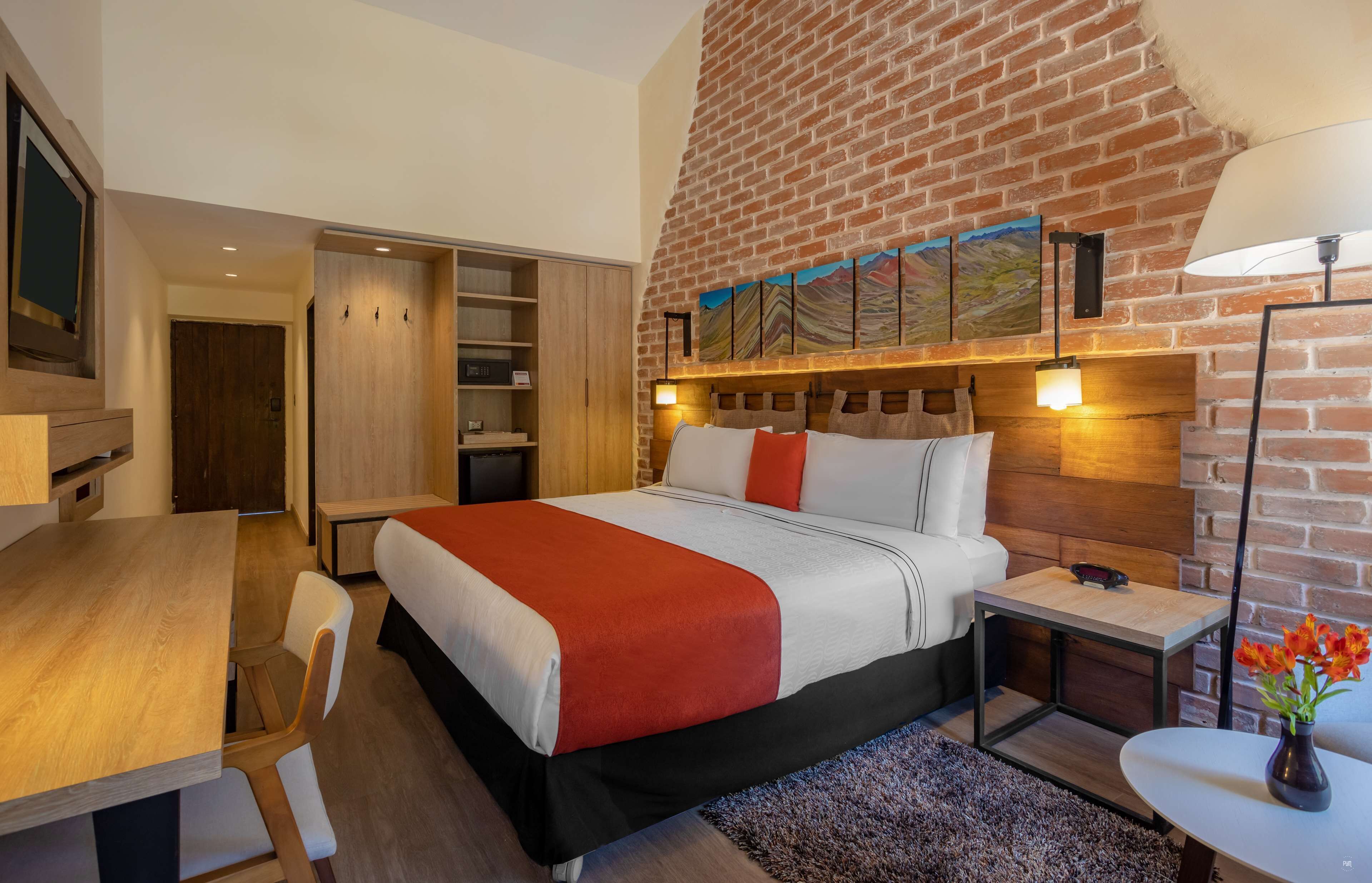 A modern hotel room with a brick wall, a large bed with a red and white bedspread, a wooden desk, a chair, a small table with a lamp, and a painting on the wall.