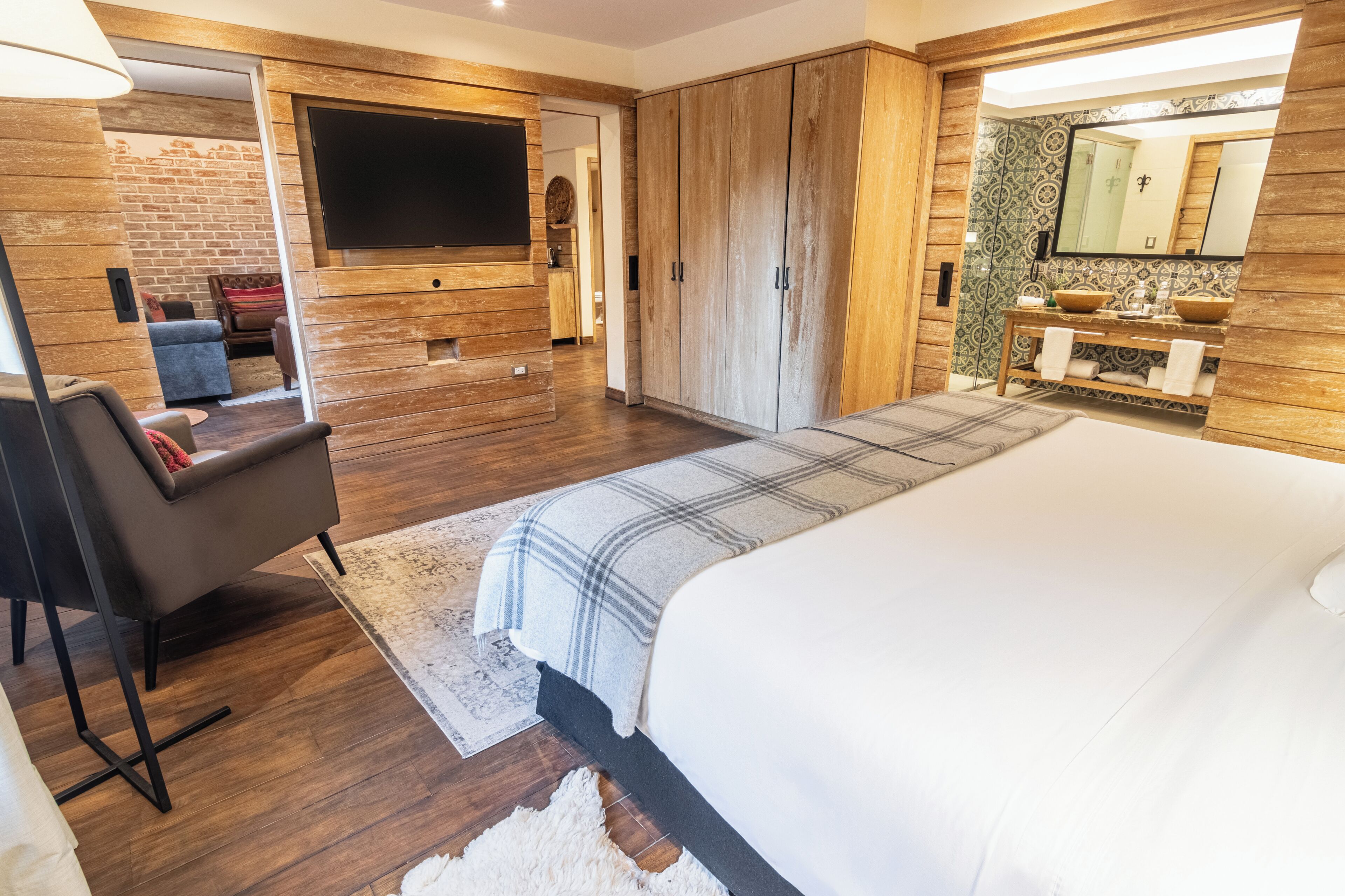 A modern bedroom with a large bed, a TV mounted on the wall, wooden wardrobe, and a bathroom with a mirror and sink.