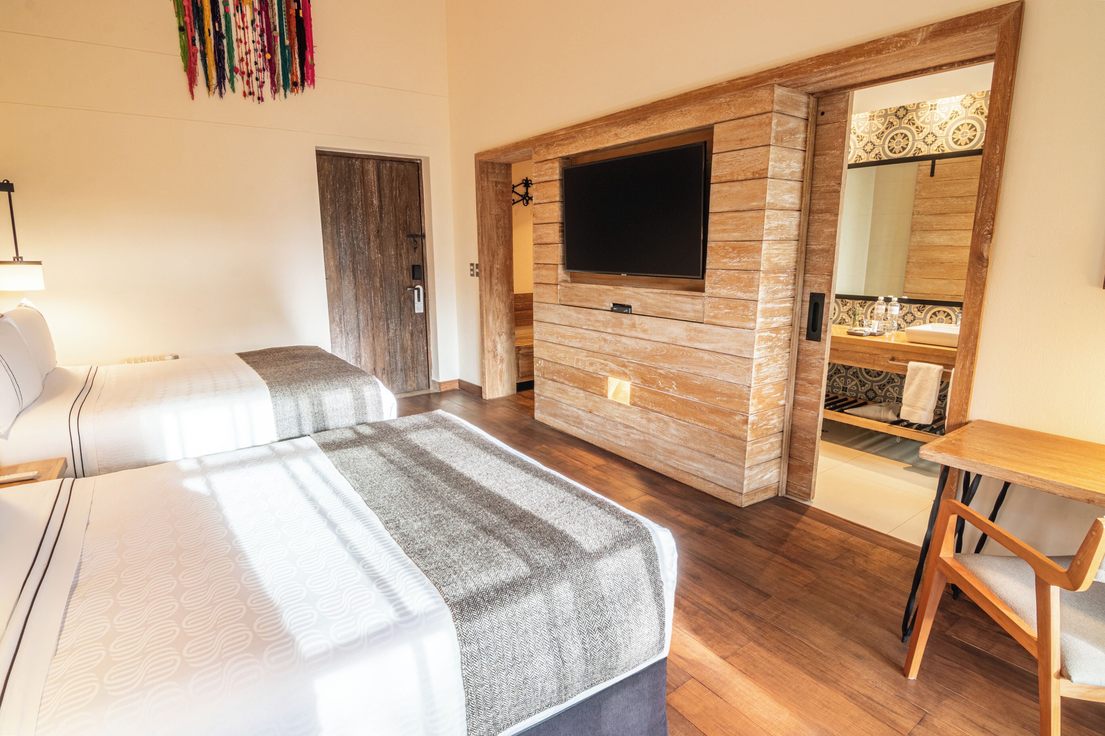 Une chambre d’hôtel avec deux lits, un téléviseur fixé à un mur en bois, un bureau en bois et une salle de bain avec un miroir et un lavabo.