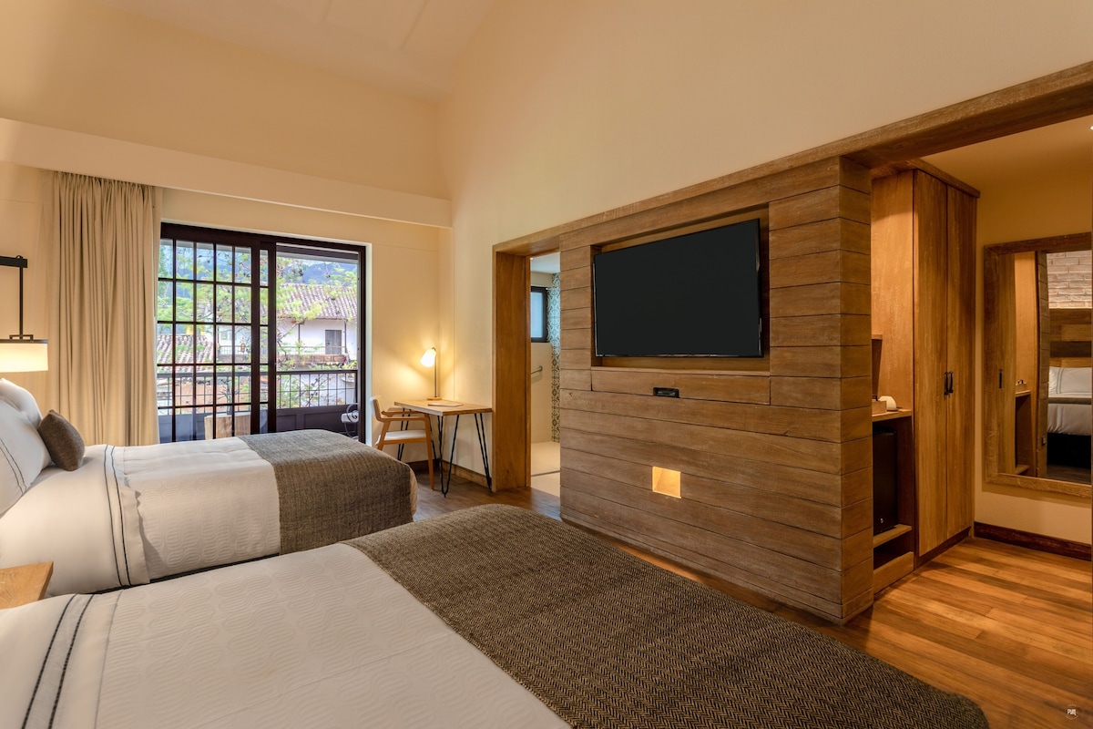 A hotel room with two beds, a television mounted on a wooden wall, a desk with a chair, and a window with a view of greenery.