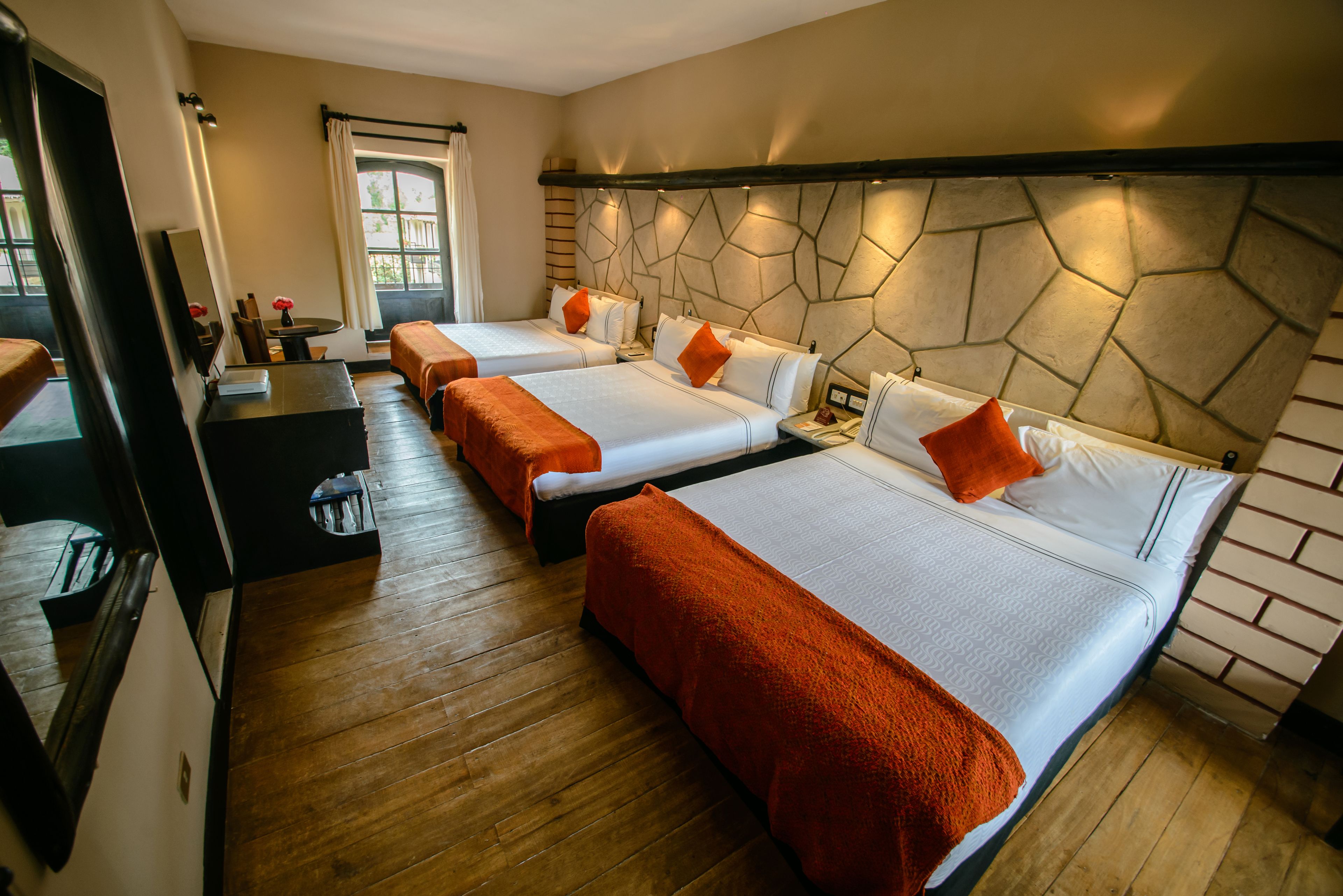 Habitación de hotel con tres camas, suelo de madera, pared decorativa de piedra y una ventana con cortinas.