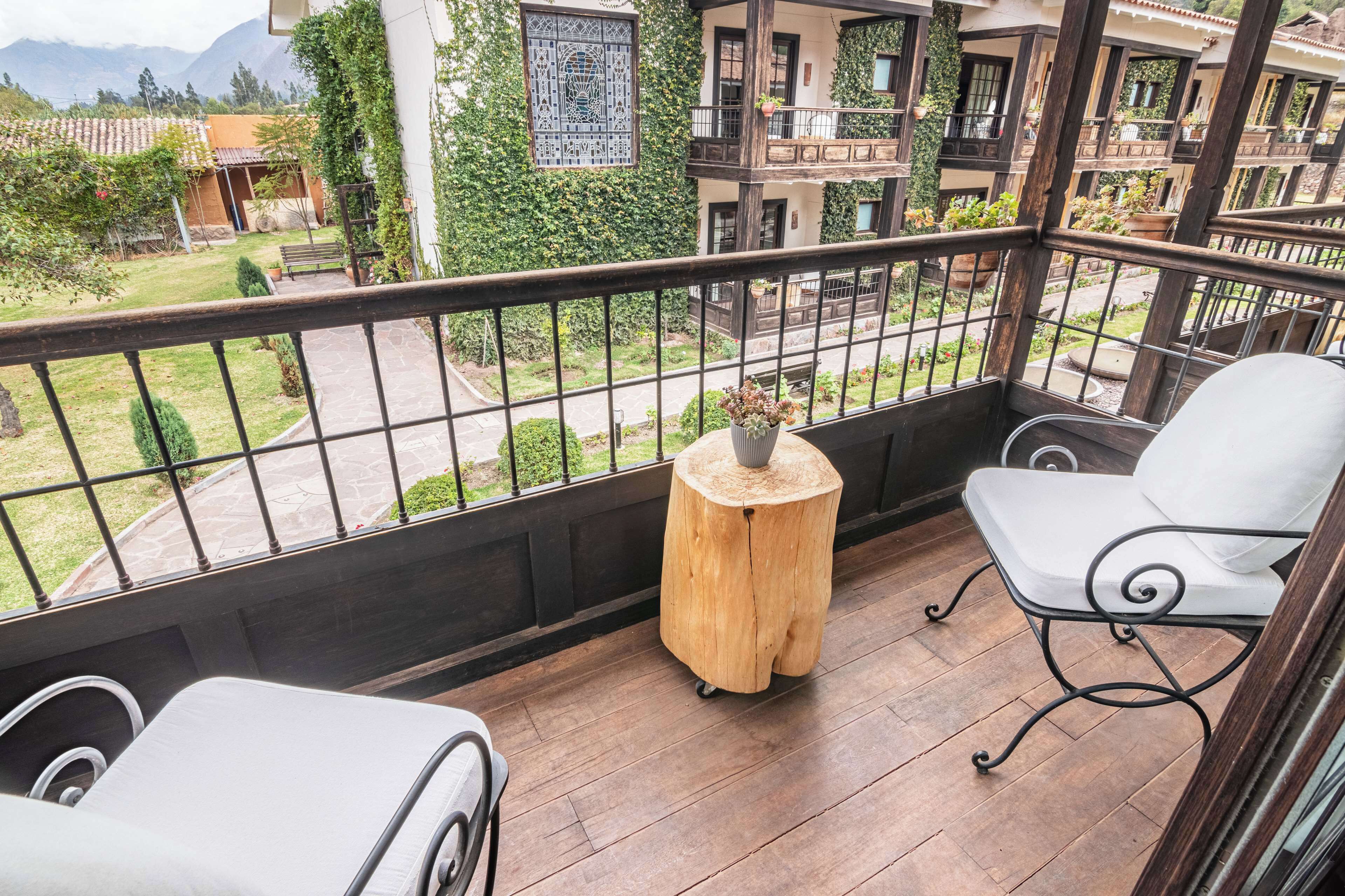 A balcony with a wooden table, a white chair, and a view of a garden and buildings.