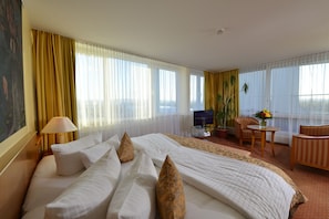 A hotel room with a large bed, a television, a desk with a computer, and a view of the city through large windows.