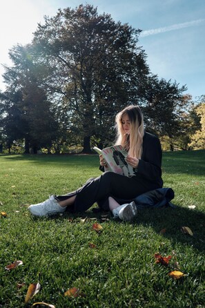 A person sitting on grass reading a book in a park with trees in the background.