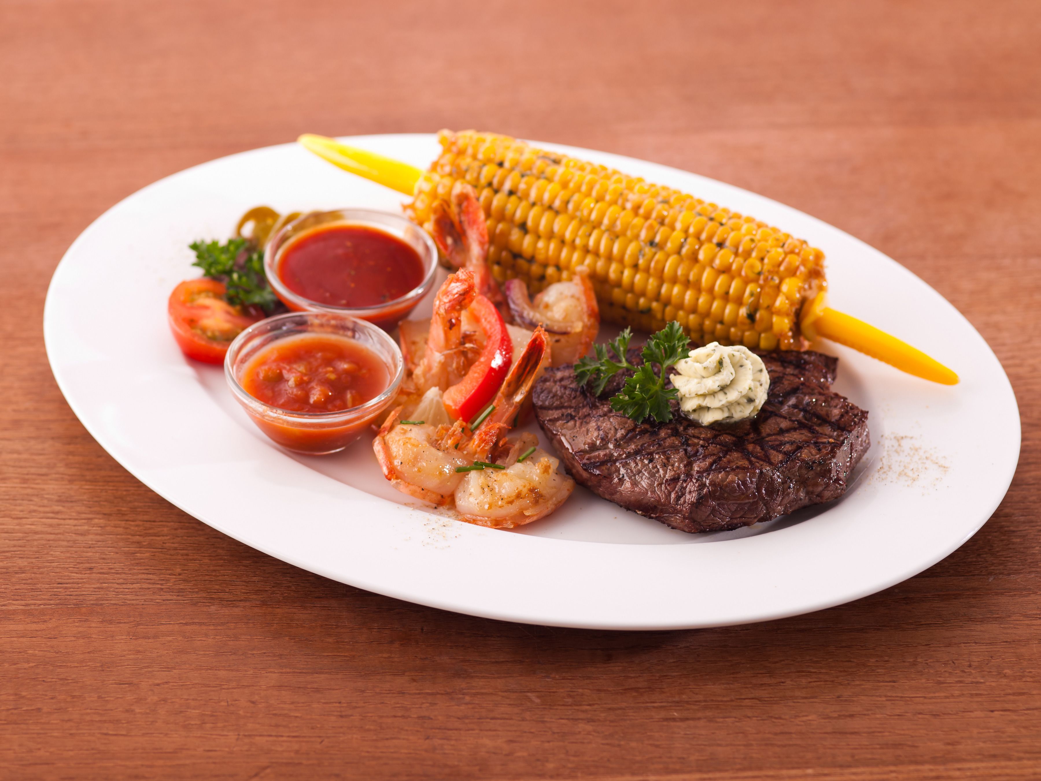 Een bord met gegrilde steak, garnalen, maiskolven en plakjes tomaat.