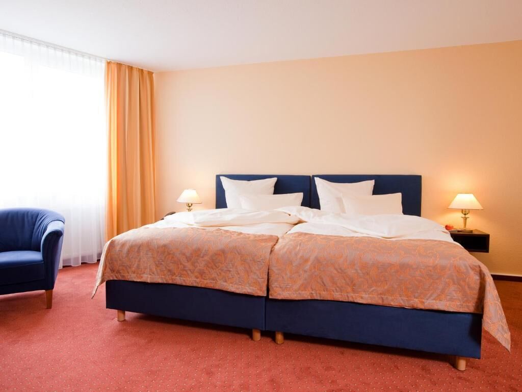 Quarto de hotel com cama de casal, duas luminárias de cabeceira, uma poltrona azul e uma janela com cortinas.