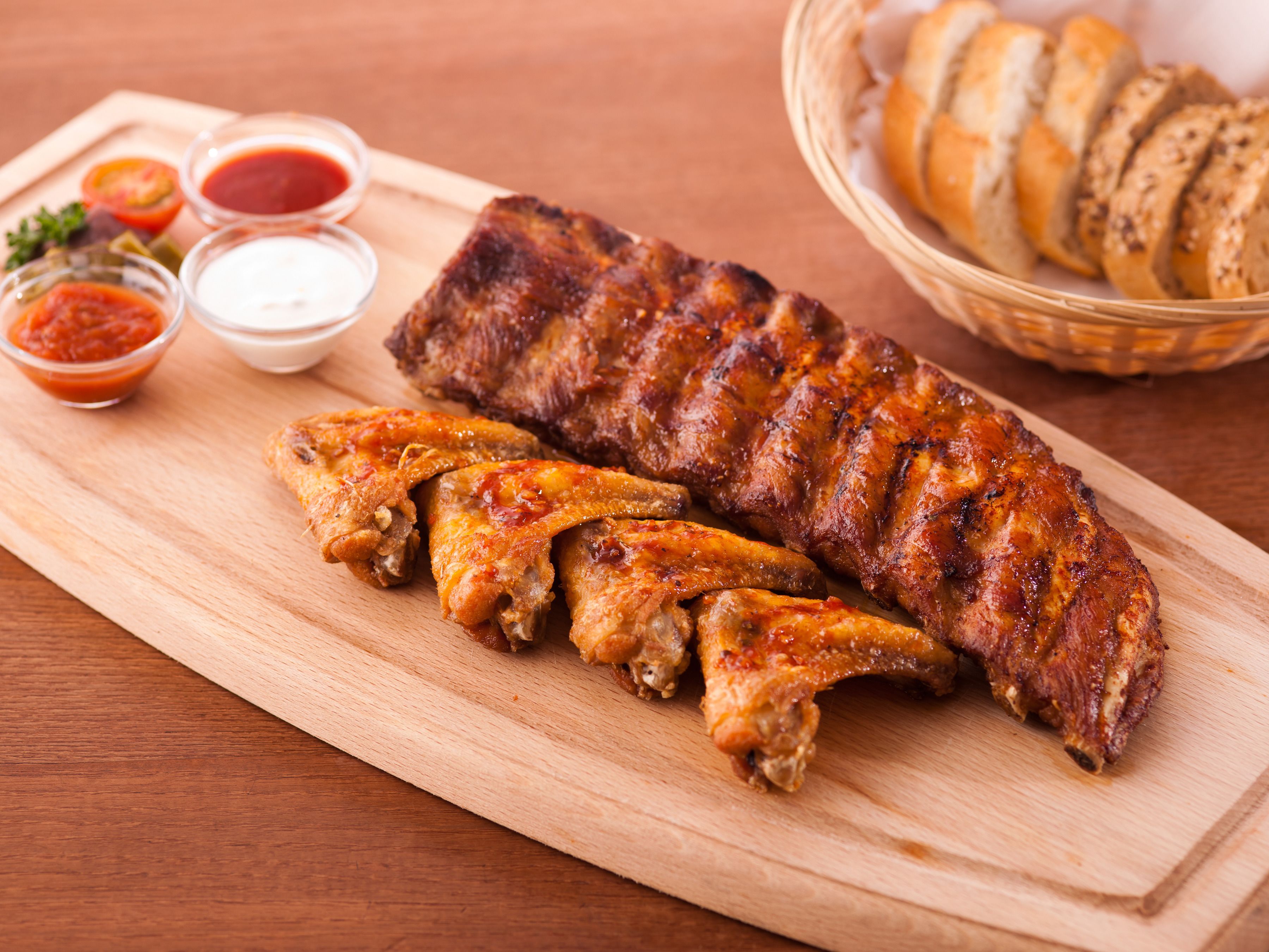 Une planche à découper en bois, sur laquelle sont disposées des travers de porc et des ailes de poulet grillées, accompagnée de sauces pour tremper et d’un panier de pain tranché.