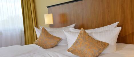 Una cama bien hecha con sábanas blancas y dos almohadas decorativas, un cabecero de madera y una lámpara de noche.