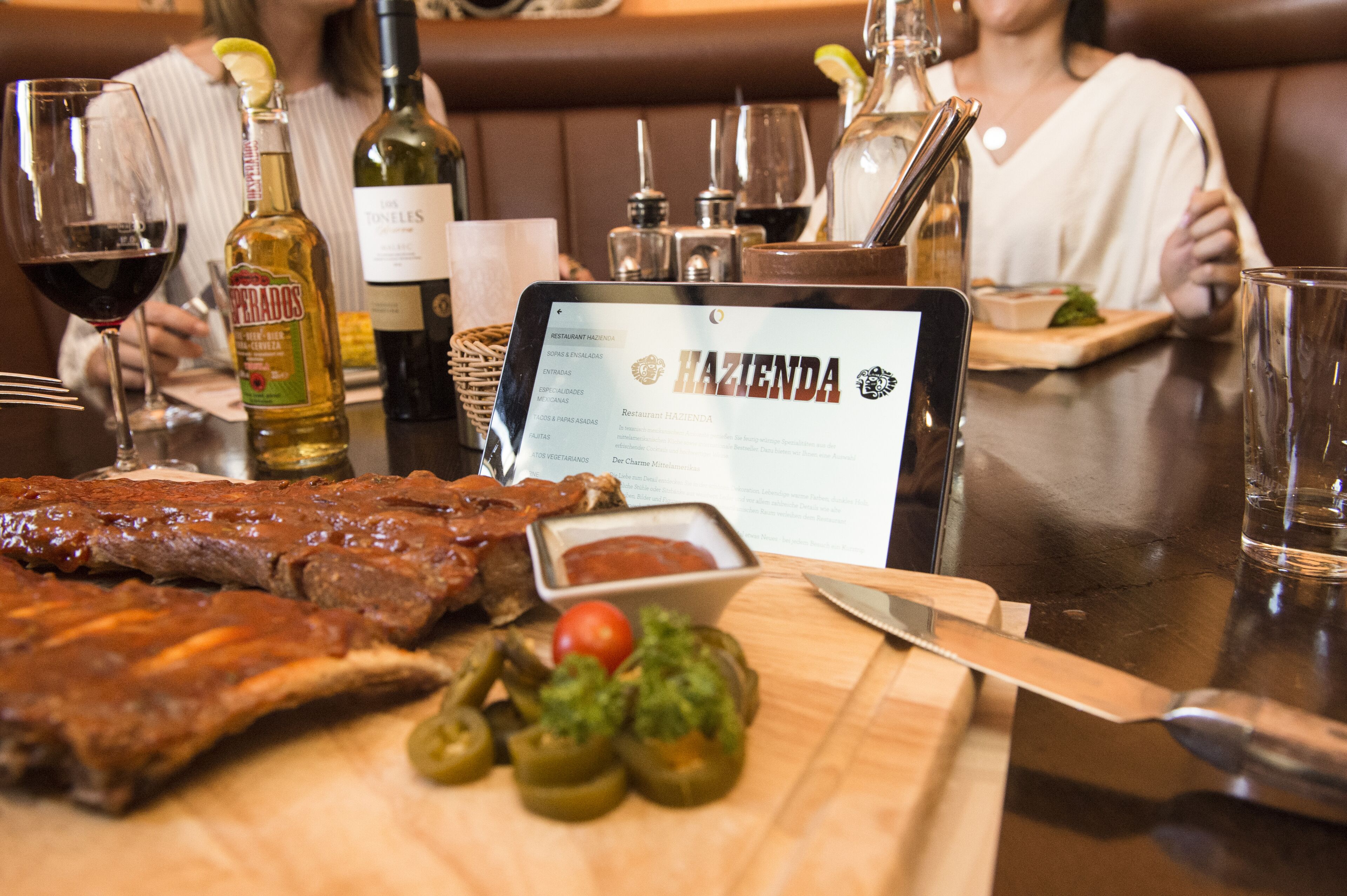 A restaurant table with a menu, a bottle of liquor, and a plate of ribs and jalapeños.