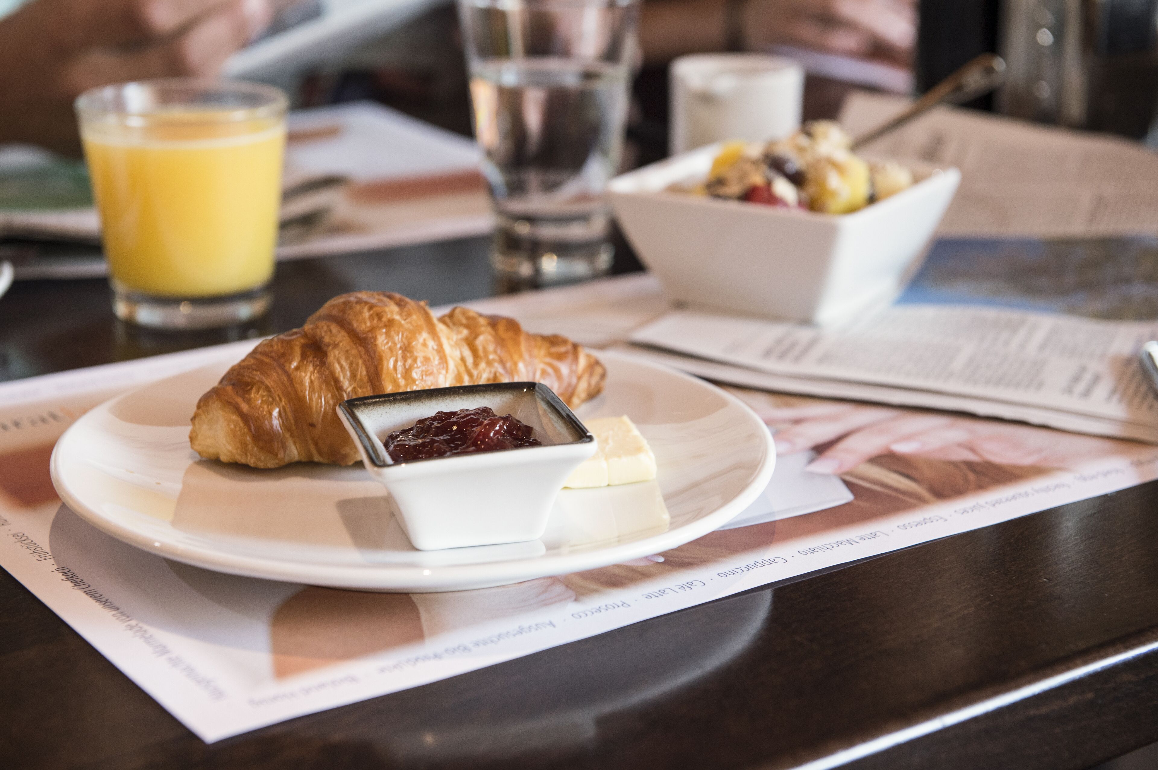 A breakfast setting with croissant, cheese, and jam on a plate, a glass of orange juice, and a bowl of fruit salad on a table.