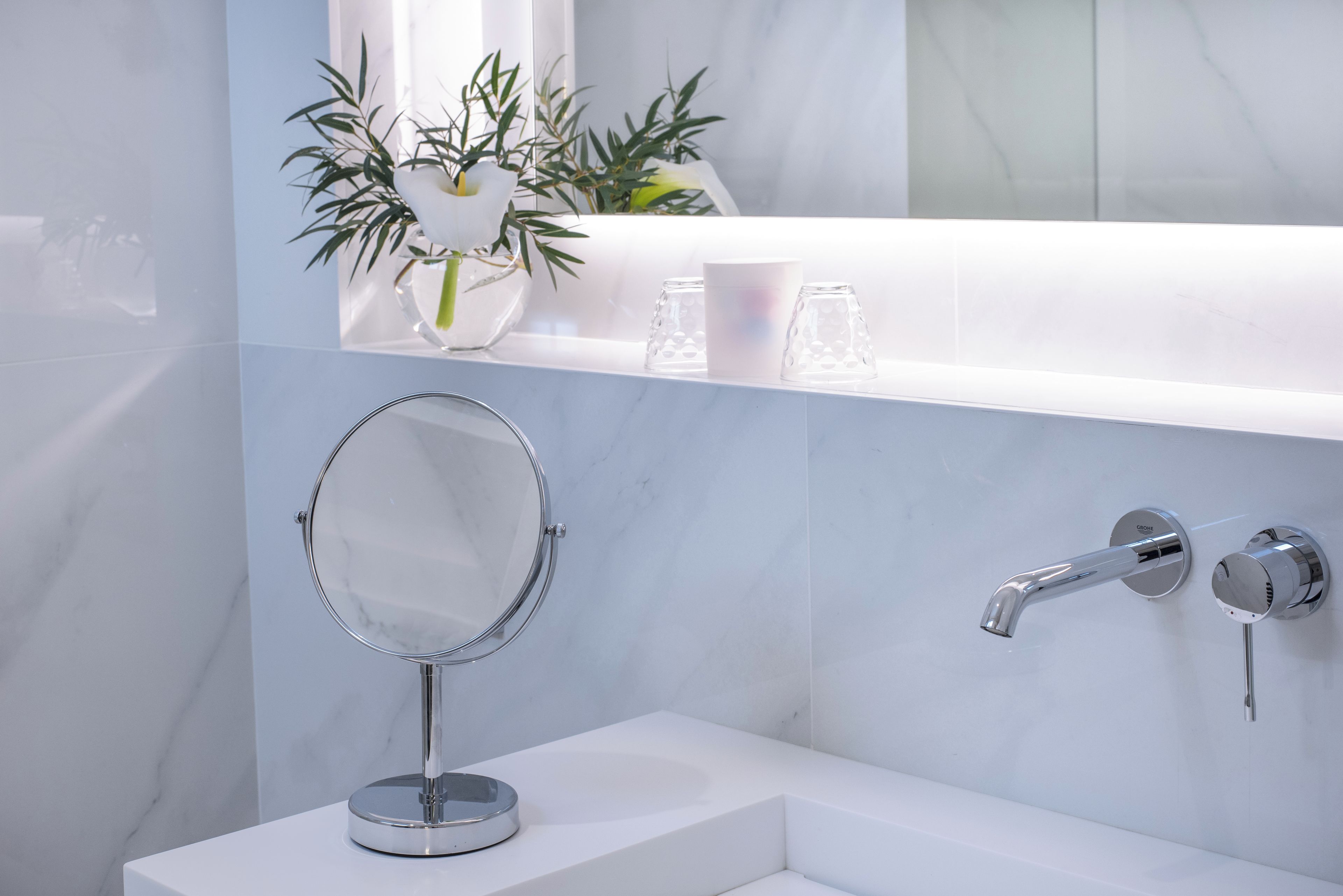 A bathroom with a marble sink, a round mirror, a faucet, and a shelf with decorative items.