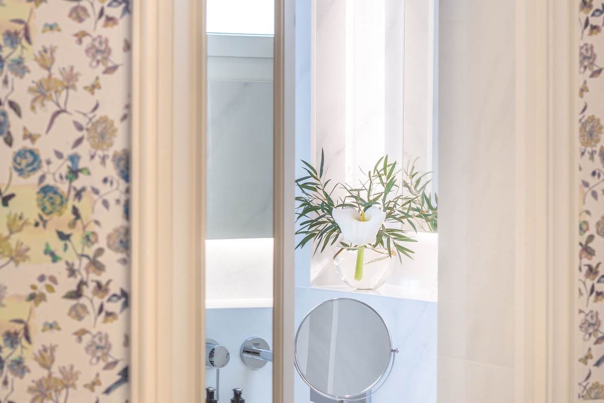 A bathroom with a floral wallpaper, a mirror, a sink, and toiletries.