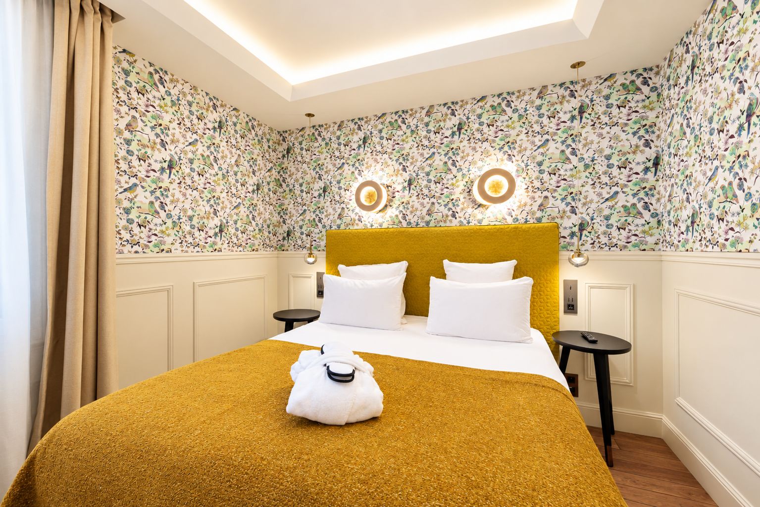 Una habitación de hotel con papel tapiz floral, una colcha color mostaza, un cabecero amarillo, dos mesitas de noche con lámparas y una toalla blanca sobre la cama.