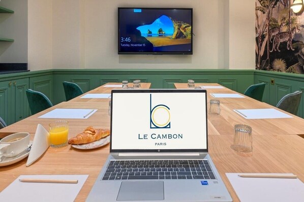 Une salle de conférence avec un ordinateur portable affichant le logo « LE CAMBON PARIS », une table en bois sur laquelle sont disposés des articles pour le petit-déjeuner, et un téléviseur fixé au mur.