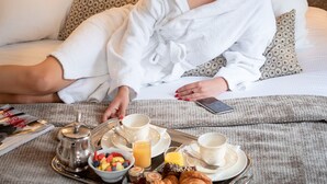A person sitting on a bed with a tray of breakfast items including croissants, fruit, and coffee. - Hotel Cambon (Paris)