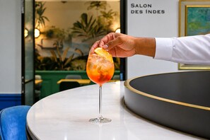 A hand holding a cocktail glass with an orange slice on a table in a room with a sign reading 'SALON DES INDES'. - Hotel Cambon (Paris)