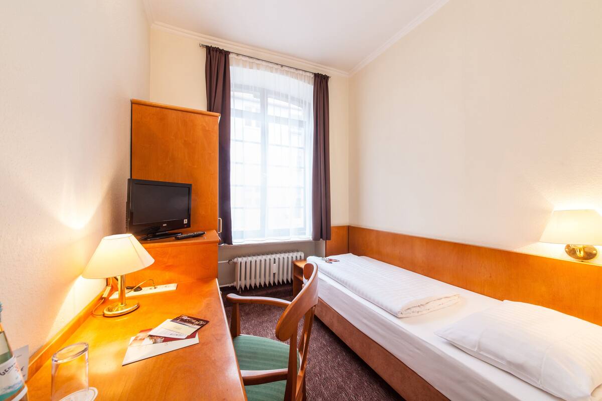 Een hotelkamer met een enkel bed, een bureau met lamp, een stoel, een televisie en een raam met gordijnen.