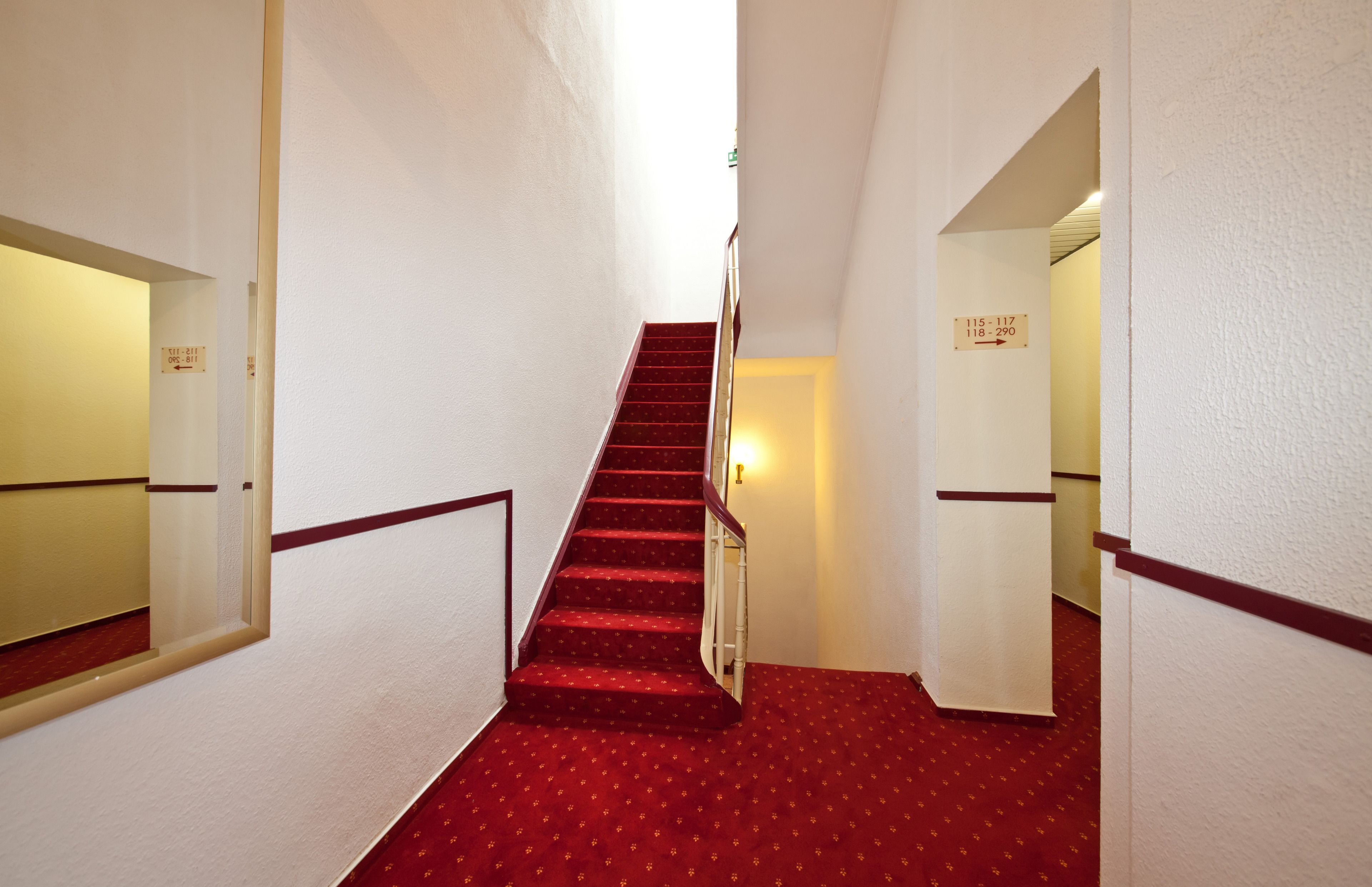 A narrow hallway with a red carpeted staircase, white walls, and a handrail.