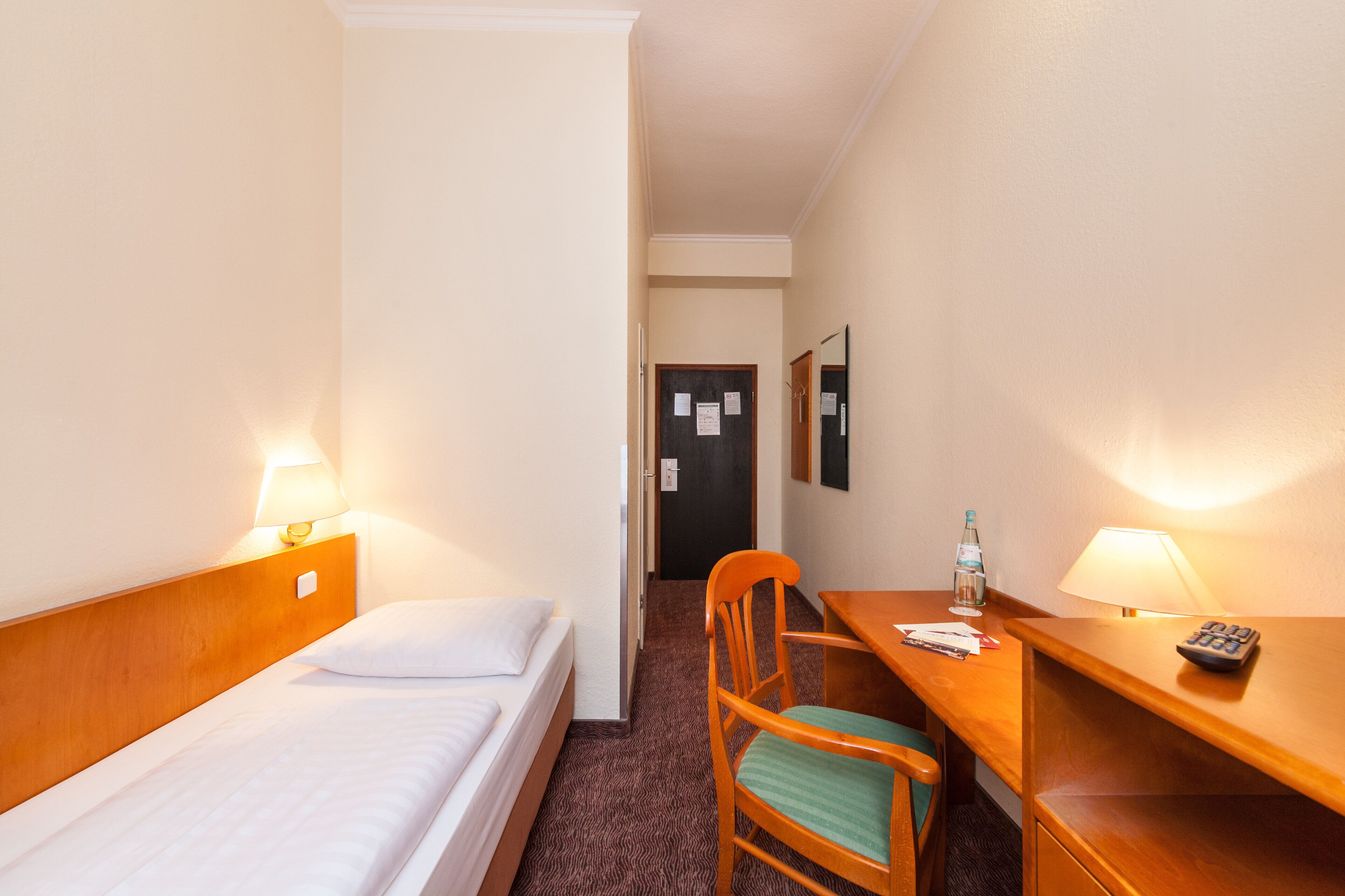 Quarto de hotel com cama, escrivaninha, cadeira, abajur e porta.