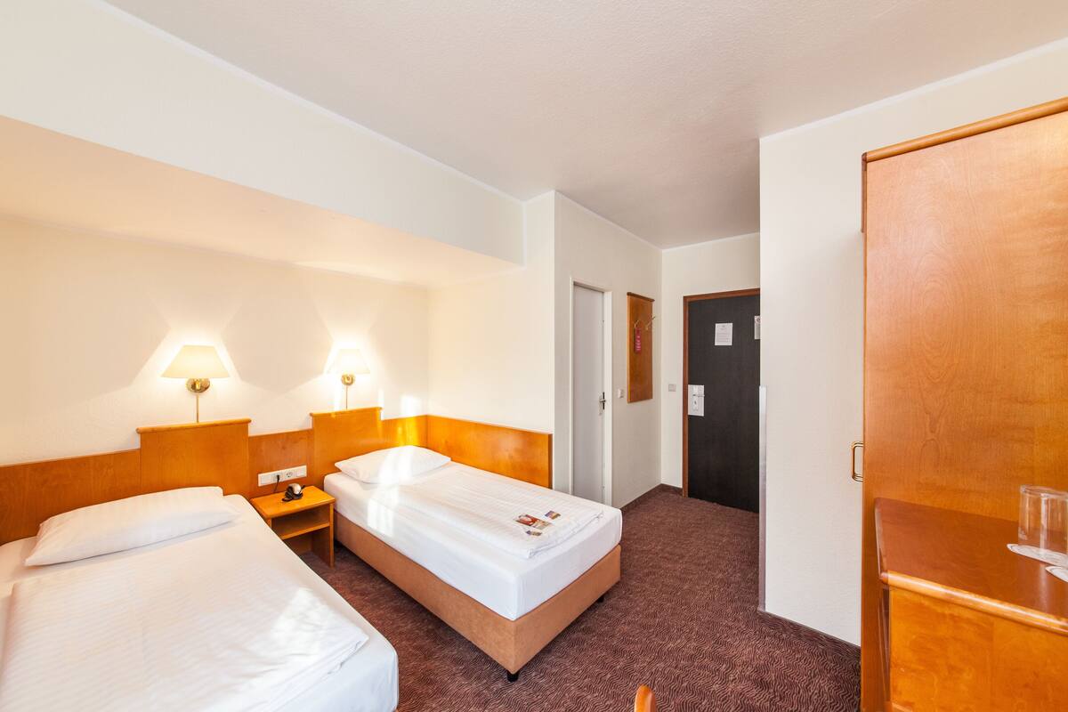 Een hotelkamer met twee eenpersoonsbedden, een houten hoofdbord, een nachtkastje, een lamp en een deur die naar een andere kamer leidt.