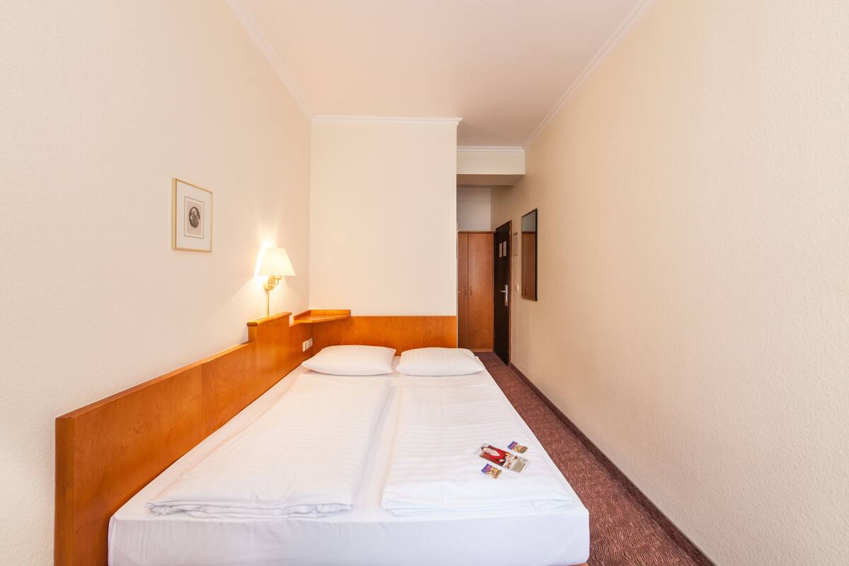 Een hotelkamer met een enkel bed, een nachtkastje met een lamp, een ingelijst schilderij aan de muur en een deur naar een andere kamer.
