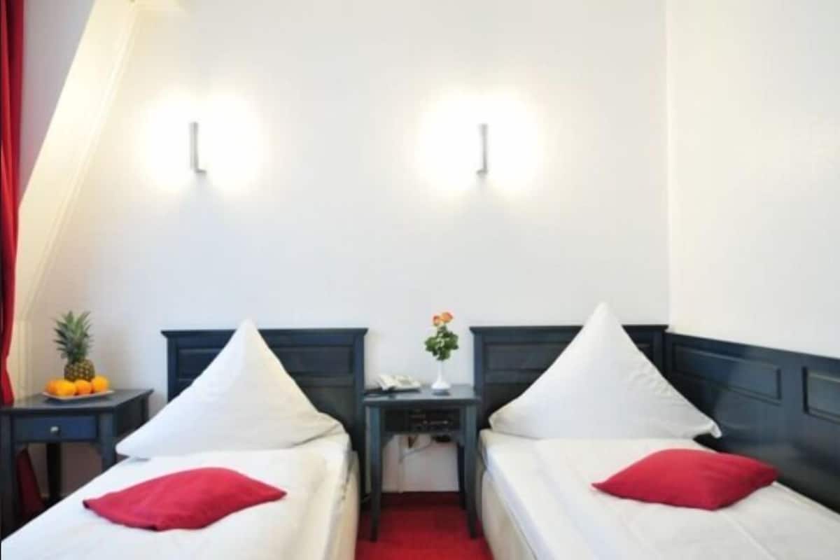 Une chambre d’hôtel avec deux lits simples, une petite table sur laquelle est posé un vase de fleurs et un rideau rouge.